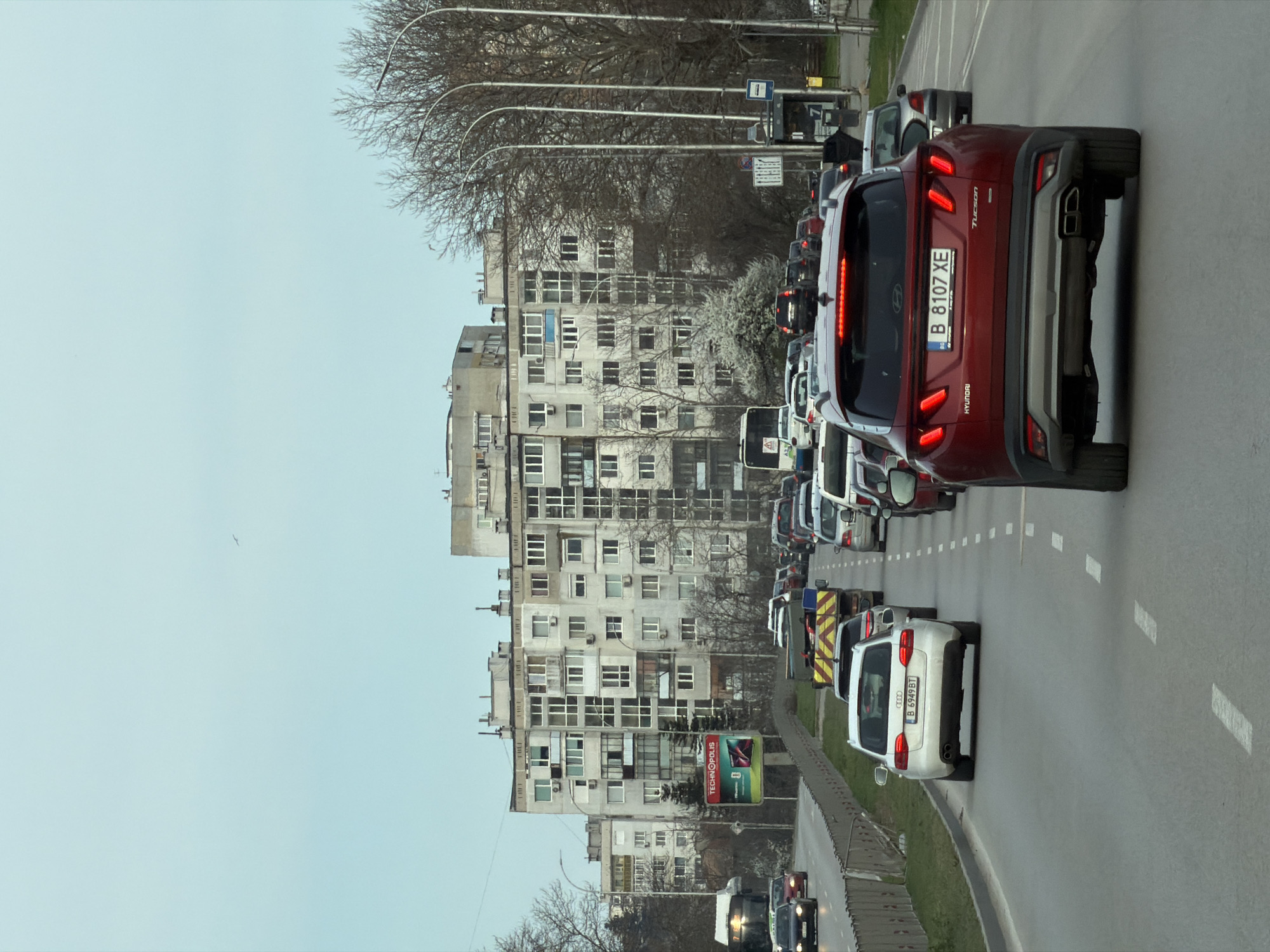 The image depicts a traffic jam in Varna, Bulgaria, with cars lined up on a multi-lane road. The camera is positioned behind a red Hyundai Tucson with the license plate B 8107 XE. A white Audi with the license plate B 6949 BT is visible in front of the Hyundai. Several buildings are in the background with bare trees and utility poles lining the street. An advertising billboard is partially visible in the lower left corner of the image. The sky is overcast, suggesting a dull or gray day. The vehicles are stationary, typical of heavy traffic congestion.