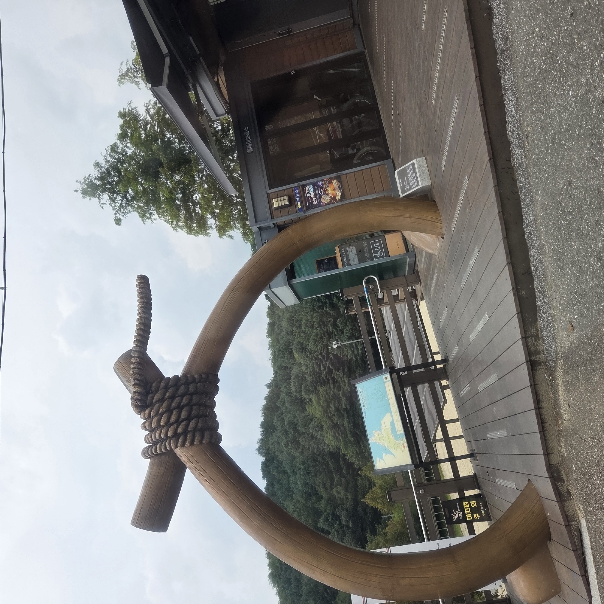 The image shows a wooden archway with a thick rope tied around its center, set on a wooden deck. To the right of the archway is a building with a wooden exterior and glass doors, possibly a visitor center or shop, with bicycles visible inside. A map is displayed on a stand to the left of the archway. The background features a dense forest covering a hillside under a cloudy sky. A sign with Korean text and a caution symbol is posted on the deck near the archway. The location is identified as Gapcheon-myeon, South Korea.