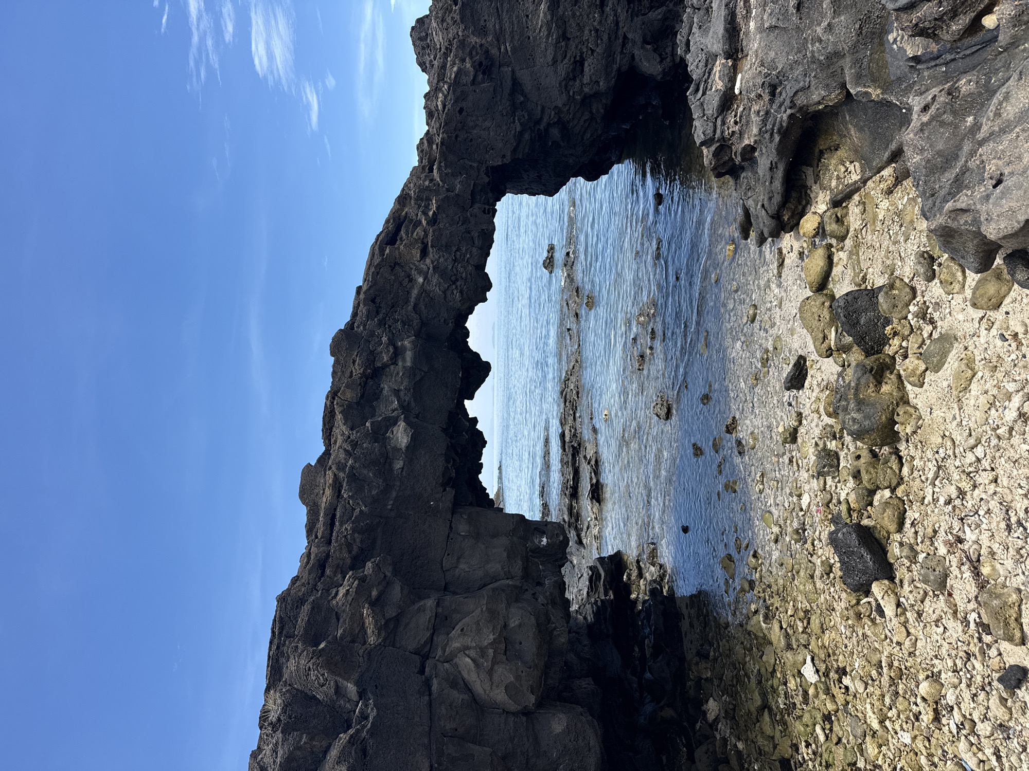 This is a vertical, eye-level, medium shot of a natural rock arch on the coast of Lý Sơn Island, Vietnam. The archway is formed from dark, jagged volcanic rock and frames a view of the calm, blue ocean and a clear sky with sparse white clouds. The foreground is a rocky beach composed of pebbles, sand, and scattered rocks. The water is clear and shallow near the shore, with some smaller rocks visible beneath the surface. The lighting suggests it is a bright, sunny day. There are no people or animals visible in the image.