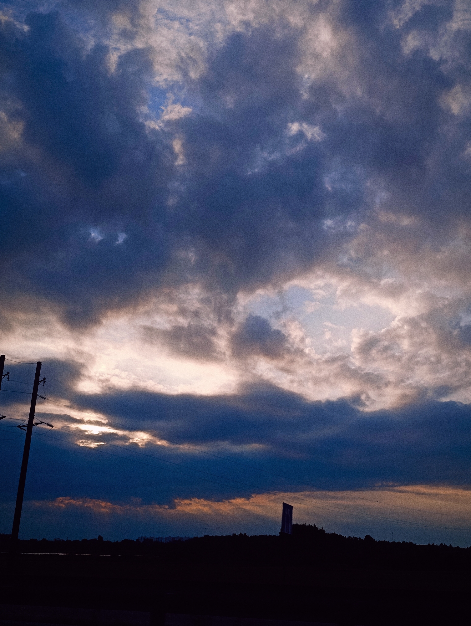 A silhouette of a dark, jagged horizon with trees and buildings is visible beneath a dramatic, cloudy sky. A tall, thin structure, possibly a monument or billboard, stands silhouetted against the horizon on the right side of the image. A utility pole with insulators and wires stands prominently on the left. The sky is filled with dark, heavy clouds with patches of lighter sky showing through, suggesting either dawn or dusk. The overall impression is one of a moody, atmospheric landscape.