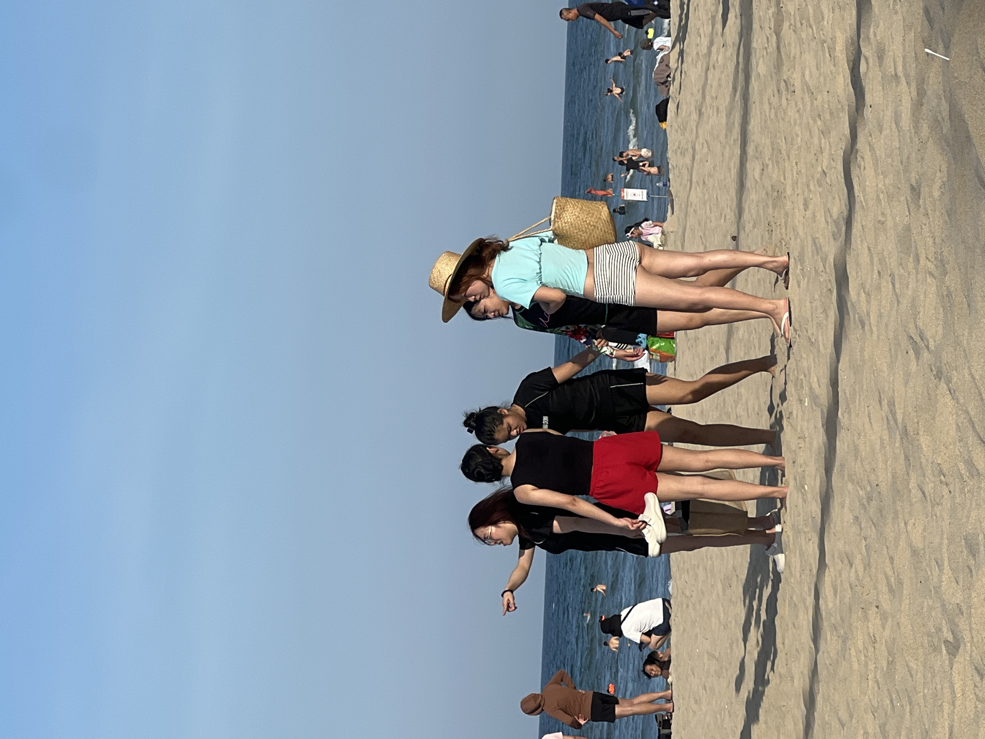 The image depicts a sunny day at a beach in Da Nang, Vietnam, with a clear blue sky and calm ocean. In the foreground, a group of five young women are gathered on the sandy beach. Two women on the left are facing away from the camera, one wearing glasses and a black shirt, while the other wears a black top and red shorts. The woman in the glasses is pointing towards the ocean. Another woman in a black shirt and black shorts is standing between them. To the right, a woman in a light blue crop top and striped shorts, wearing a straw hat, is looking towards the ocean. Next to her, another woman in a black and floral rash guard and black shorts is also present. They appear to be casually conversing.
In the background, numerous people are visible enjoying the ocean, swimming, and wading in the water. Some are closer to the shore, while others are further out. The beach is populated with beachgoers of various ages. The overall atmosphere suggests a leisurely day at the beach.