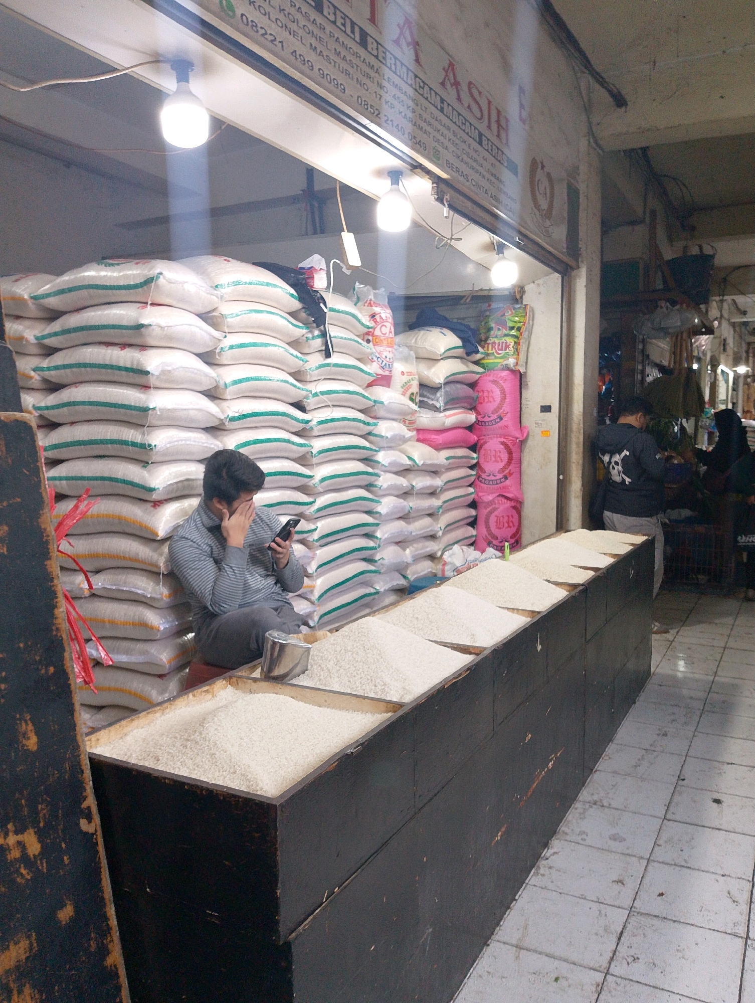 The image depicts a street-level scene at a rice stall in Lembang, Indonesia. A man wearing a striped long-sleeved shirt and dark pants is seated on the floor, leaning against large sacks of rice, and looking at his mobile phone. In front of him, there is a wooden counter filled with loose rice. The stall is lined with numerous large sacks of rice, some with green and yellow stripes. Behind the man, a sign is partially visible with text that appears to be in Indonesian. To the right of the stall, two people are visible in the background, one wearing a black jacket with a skull graphic and the other in a dark hijab. The environment appears to be an indoor market or a covered street stall, with tiled flooring. The lighting is somewhat dim, with bright lights visible above the stall, illuminating the sacks of rice.