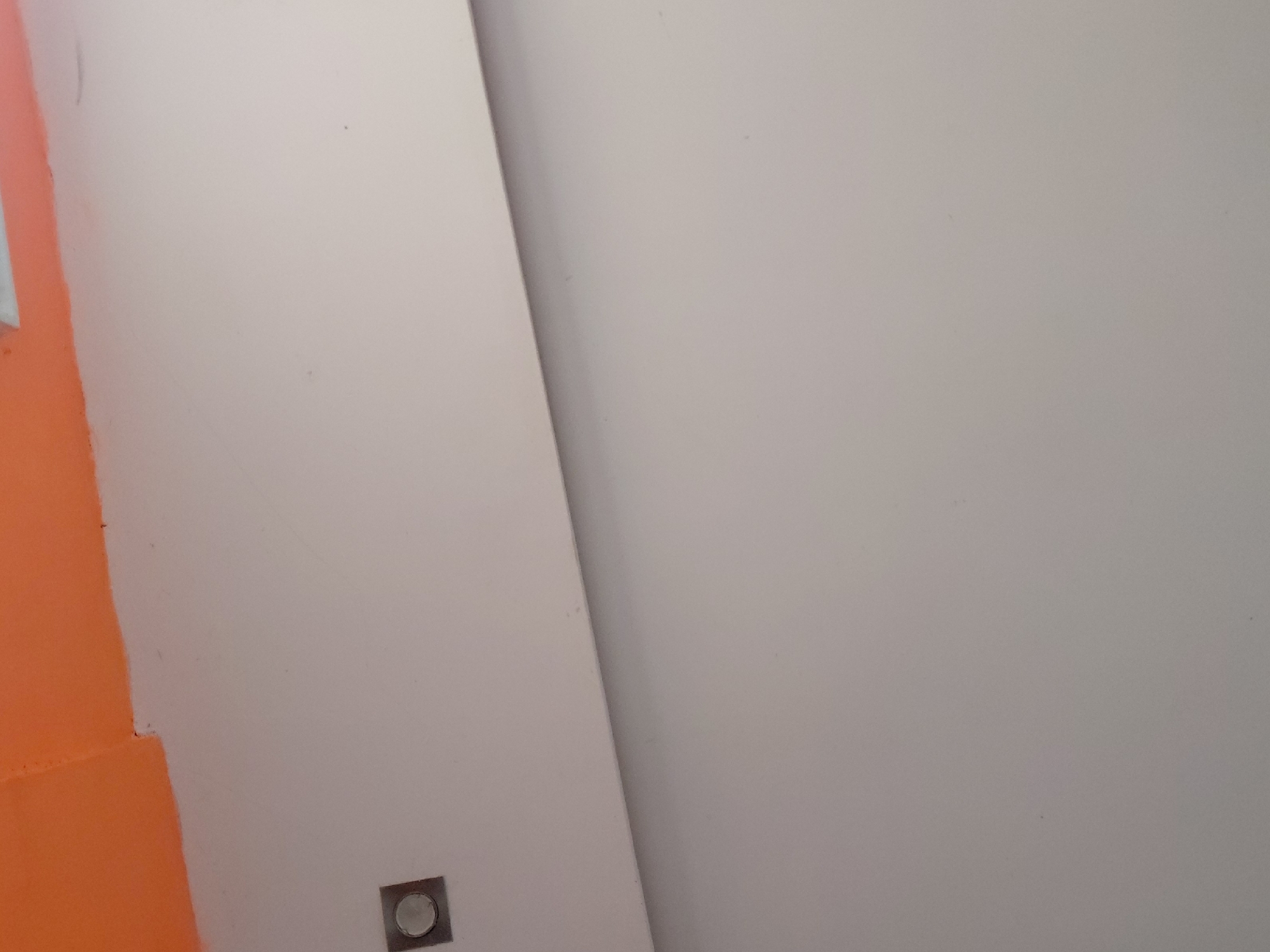 This image presents a close-up, static view of an interior architectural detail in Santa Ana, Venezuela. The scene is dominated by a large, smooth surface painted in a light gray or off-white hue, which covers the majority of the frame, especially on the right side. On the far left, a distinct vertical section of a wall or panel in a vibrant orange color is visible, creating a strong contrast.
A sharp, diagonal line runs from the upper center towards the lower center, dividing the light gray surface into two planes, suggesting an interior corner, an architectural column, or a transition between two different wall sections. The rightmost portion of the light gray area appears slightly darker, possibly indicating a subtle shadow or a different angle of incidence for the ambient light.
At the bottom-center, on the light gray wall, there is a small, square, metallic fixture with a circular depression or button in its center, resembling an electrical outlet or a light switch. The wall surfaces appear generally clean with minor speckles or dust particles visible upon closer inspection. There are no people, explicit activities, or discernible outdoor elements in the frame. The lighting is soft and diffused, offering no clues about the time of day or external weather conditions. No text is visible.