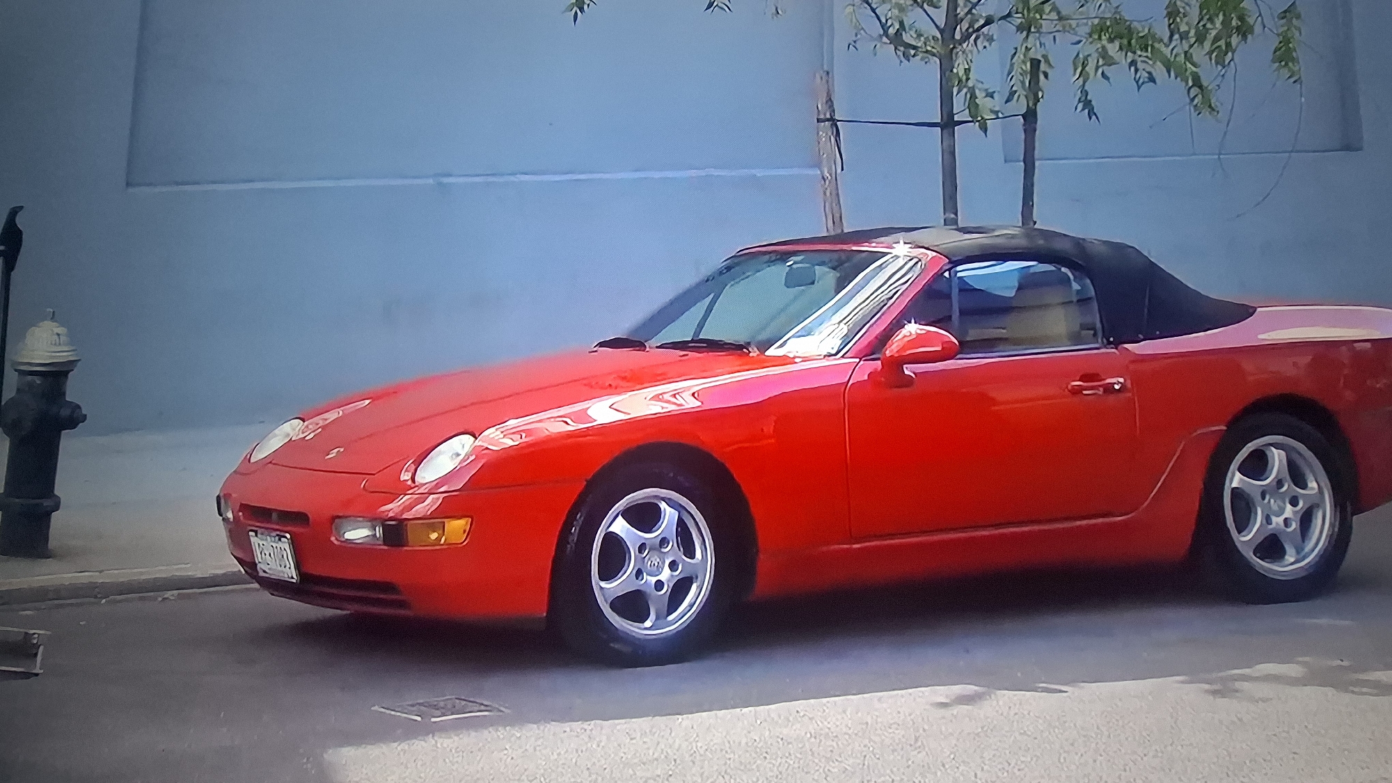 A red Porsche 968 convertible is parked on a city street. A black fire hydrant is to the left of the vehicle. A tree is to the right, and a light blue wall serves as the background. The location is Unknown, Unknown.