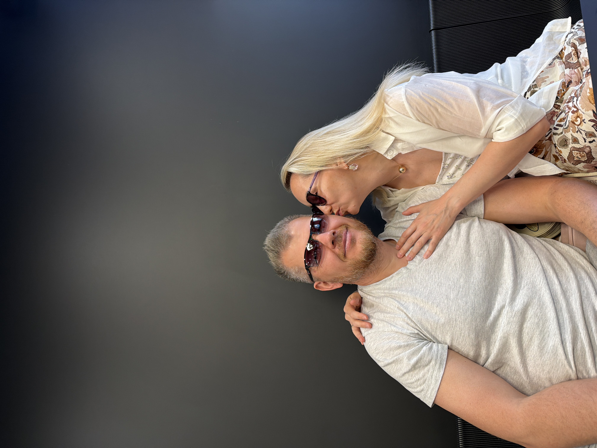A medium shot depicts a fair-skinned woman kissing a fair-skinned man on the forehead; both wear sunglasses. The man is reclining, with the woman leaning over him, her left arm around his neck and her right hand on his chest. The woman has long blonde hair and wears a white shirt over a floral-print top and skirt. The man has short, graying hair and a beard and wears a gray t-shirt. The background is a solid, dark color. The location is not evident from this image. The image does not feature the city of Izhevsk, Russia.