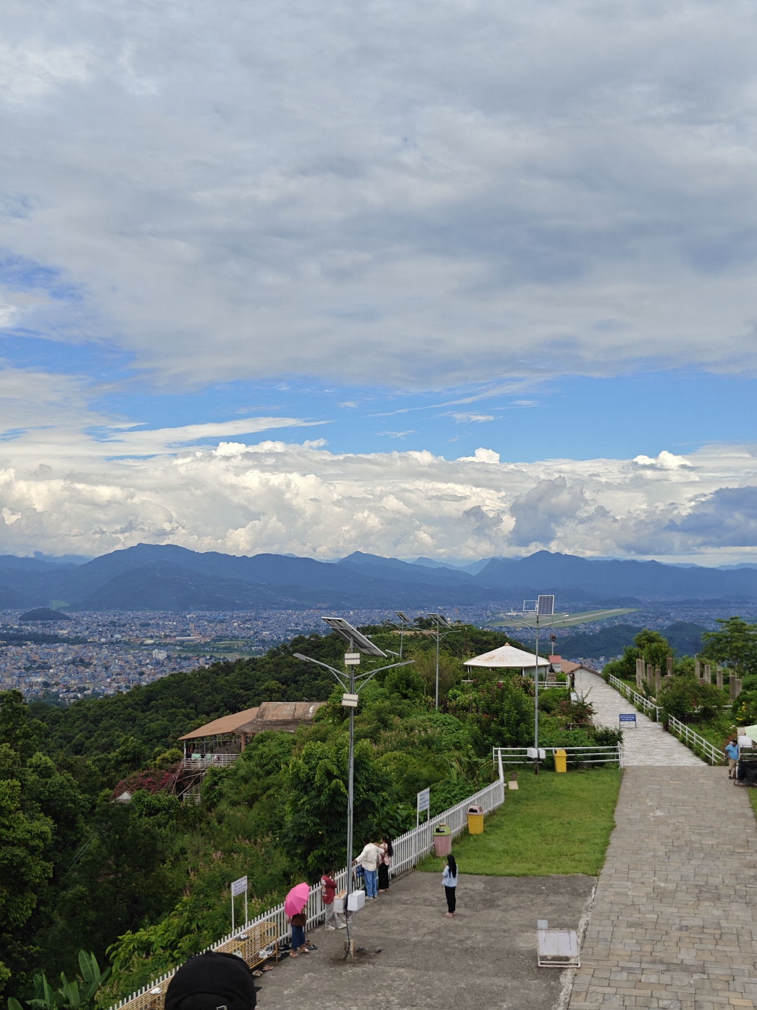 The image depicts a scenic view from a hilltop overlooking the city of Pokhara, Nepal. In the foreground, a paved pathway leads down a gentle slope, bordered by a white railing. Several people are visible on the pathway, some standing and observing the view, while others are walking. One person holds a pink umbrella. To the left of the pathway, lush green trees and vegetation cover the hillside. A white building with a conical roof and a shaded patio is nestled among the greenery. Further down the hillside, more buildings and the dense urban landscape of Pokhara extend towards a range of blue mountains in the distance. The sky is filled with a mix of white, fluffy clouds and patches of blue. There are also several lampposts with solar panels along the pathway, indicating a maintained tourist or recreational area. In the far distance, an airport runway is visible.