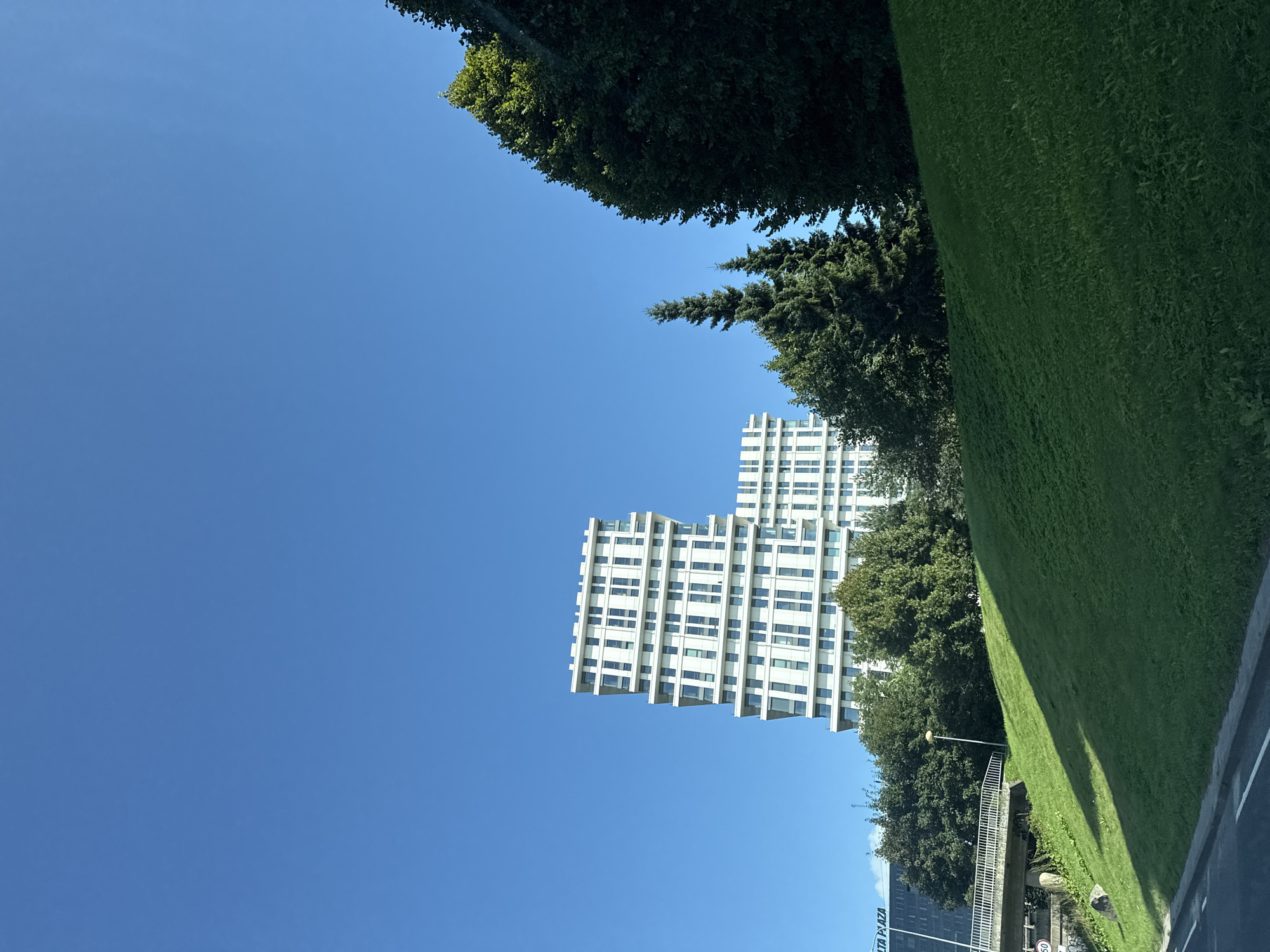 The image shows a modern white apartment building with a distinctive stepped design, located in the city of Tallinn, Estonia. The building is situated behind a grassy hill, and a bridge can be seen on the left side of the frame. Tall evergreen trees frame the right side of the building, and a clear blue sky occupies the upper portion of the image. The building appears to be part of a larger development, with another similar structure visible behind it. The overall scene is one of urban architecture integrated with natural landscaping.