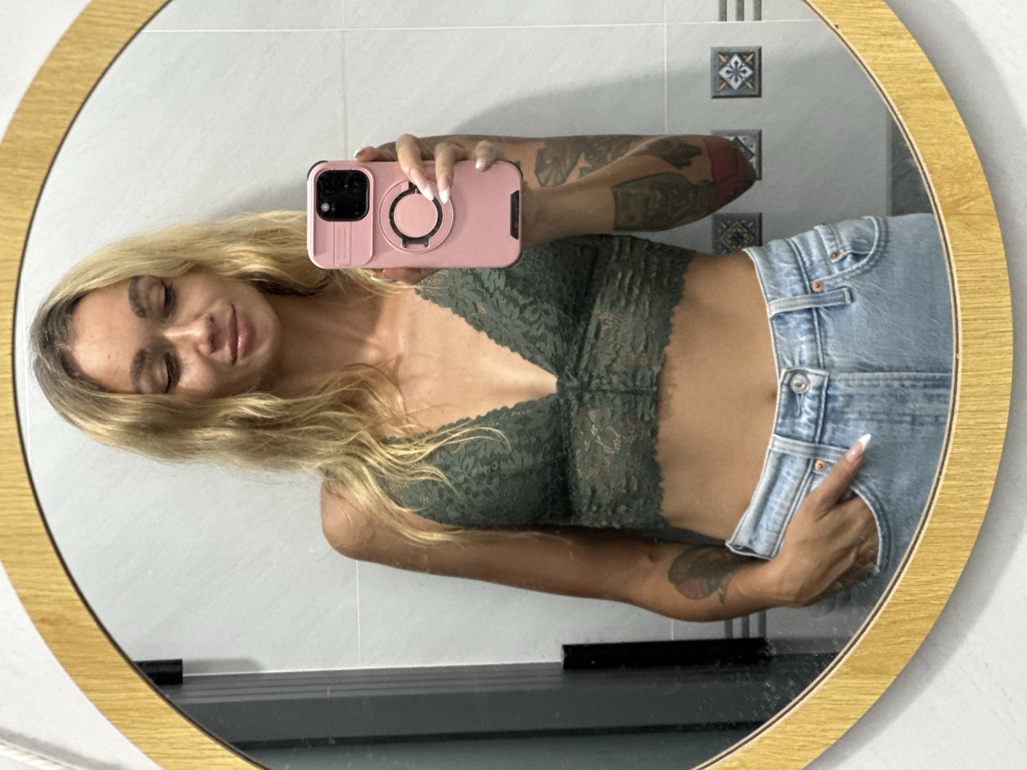 A woman with blonde hair and tattoos on her arms is taking a mirror selfie in a bathroom. She is wearing a green lace bralette and light blue jeans. She is holding a pink phone in her right hand. The bathroom has white tiled walls and a round mirror with a wooden frame. The image is a reflection in the mirror. The location is the city of Mũi Né, Vietnam.
