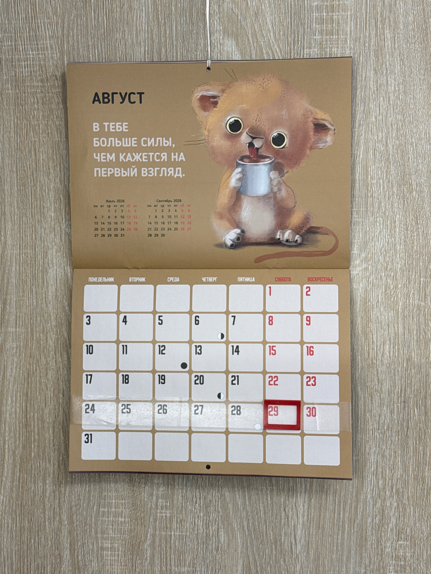 The image shows a wall calendar for August. The calendar features a cartoon illustration of a lion cub drinking from a mug. The text on the calendar is in Russian and reads "August" and "There is more strength in you than it seems at first glance." The calendar also shows a grid for August 2026, with days of the week listed at the top: Monday, Tuesday, Wednesday, Thursday, Friday, Saturday, Sunday. The image does not provide any information about the location of Lyubertsy, Russia.