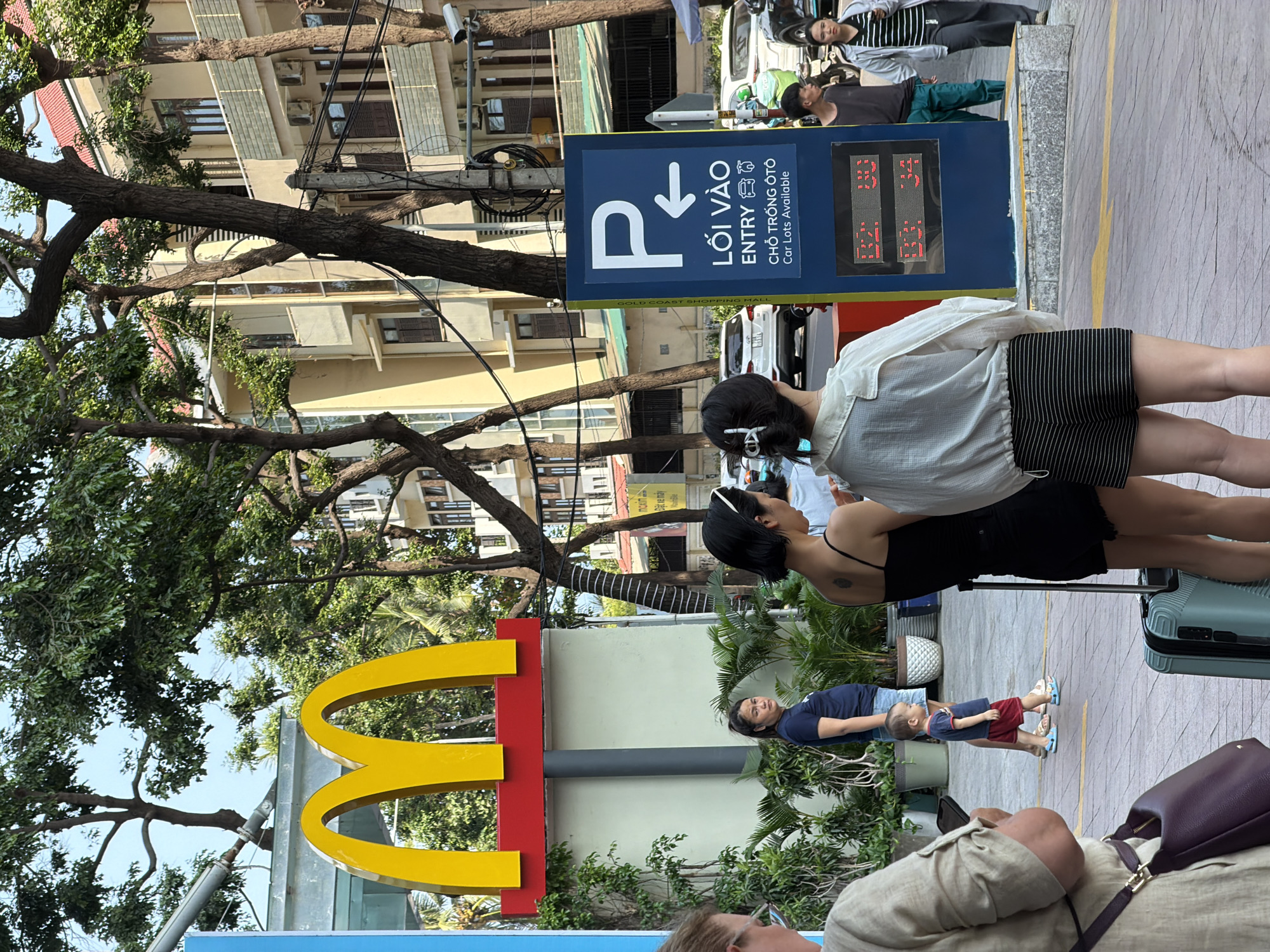 The scene is in Nha Trang Ward, Vietnam. A blue parking sign with the words "LOI VAO ENTRY" and "CHO TRONG OTO Car Lots Available" is visible near a McDonald's sign. Two women stand beside the parking sign, one wearing a white shirt and striped shorts, the other in a black dress. A child in a blue shirt and red shorts stands nearby. A suitcase sits on the ground beside the women. The background features a building and a tree with dense foliage. Overhead power lines are also present. The scene appears to be a busy street. Other pedestrians are visible behind the parking sign.