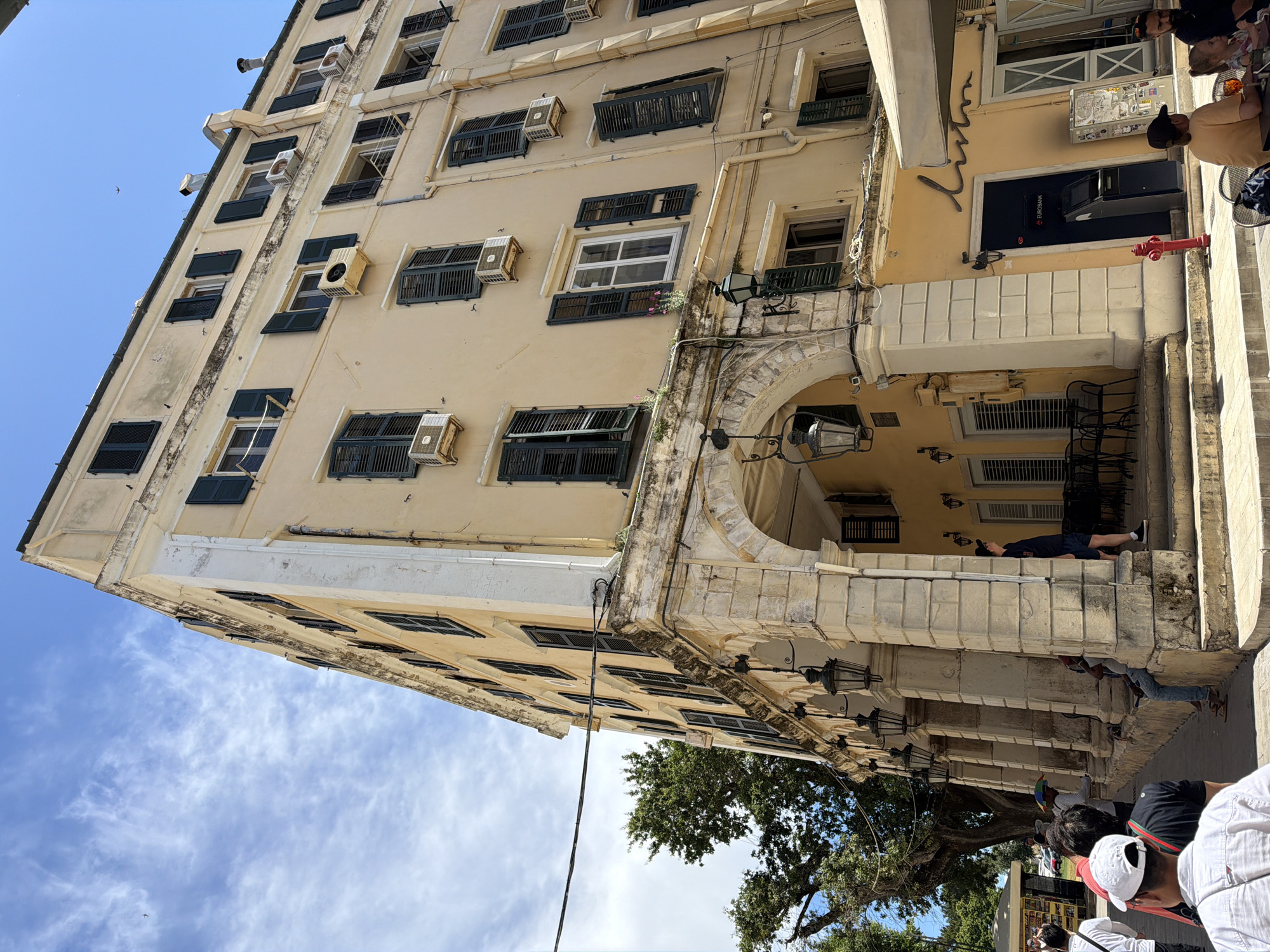 This image captures a vibrant street scene in Corfu, Greece, focusing on a multi-story building under a bright, clear blue sky. The building's facade is painted a light yellow, featuring numerous windows fitted with traditional dark green wooden shutters, many of which are open. Several white air conditioning units are mounted beneath the windows.
At street level, the building is supported by decorative stone archways and columns, beneath which various activities unfold. In the foreground, a person wearing a dark shirt and cap is casually lying on a stone ledge, resting. Further along, other individuals are seated at outdoor tables, enjoying a cafe or restaurant. A large, green-leafed tree is visible in the lower left, while a small bird soars in the distant sky.
The scene is bathed in daylight, casting shadows that indicate a sunny day with only a few wispy clouds. Notable details include ornate black street lamps affixed to the stone structure, a red fire hydrant, and an ATM integrated into the building's facade. The ATM prominently displays the text "EUROBANK," and above it, a stylized cursive logo, "λυμ," is visible. The overall atmosphere is relaxed and characteristic of a lively Mediterranean town.