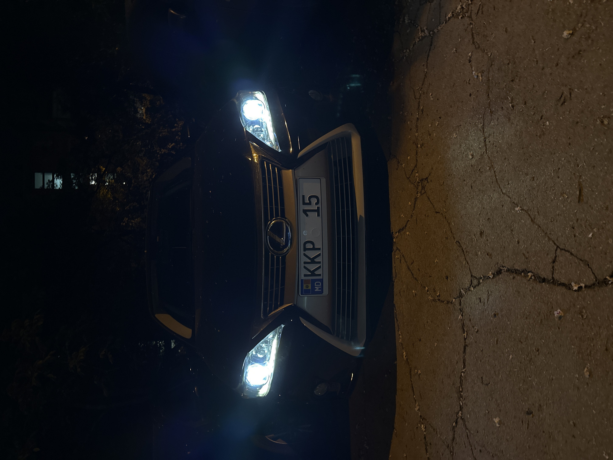 A Lexus automobile is parked in Unknown, Unknown. The car's license plate reads "MD KKP 615". The car's headlights are on. The car is parked on a cracked surface. The surroundings are dark.