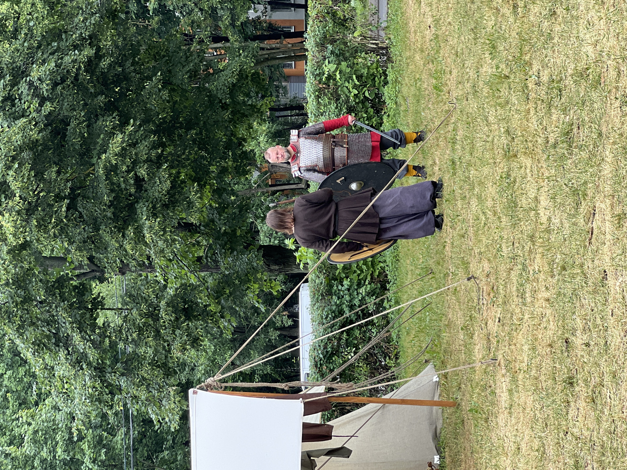 Two individuals dressed in historical reenactment attire, likely representing Vikings or similar medieval warriors, stand on a grassy field. The person on the left wears a dark brown tunic and loose-fitting grey trousers, holding a round shield. The person on the right is clad in a chainmail-style vest over a red tunic and black trousers with yellow leg wraps, holding a sword. A canvas tent is visible to the left, supported by wooden poles and ropes. In the background, dense green trees line the perimeter of the field, with a hedge and a building visible beyond them. The scene appears to be an outdoor event, possibly a historical reenactment festival, set within a park or open space in Moscow, Russia.