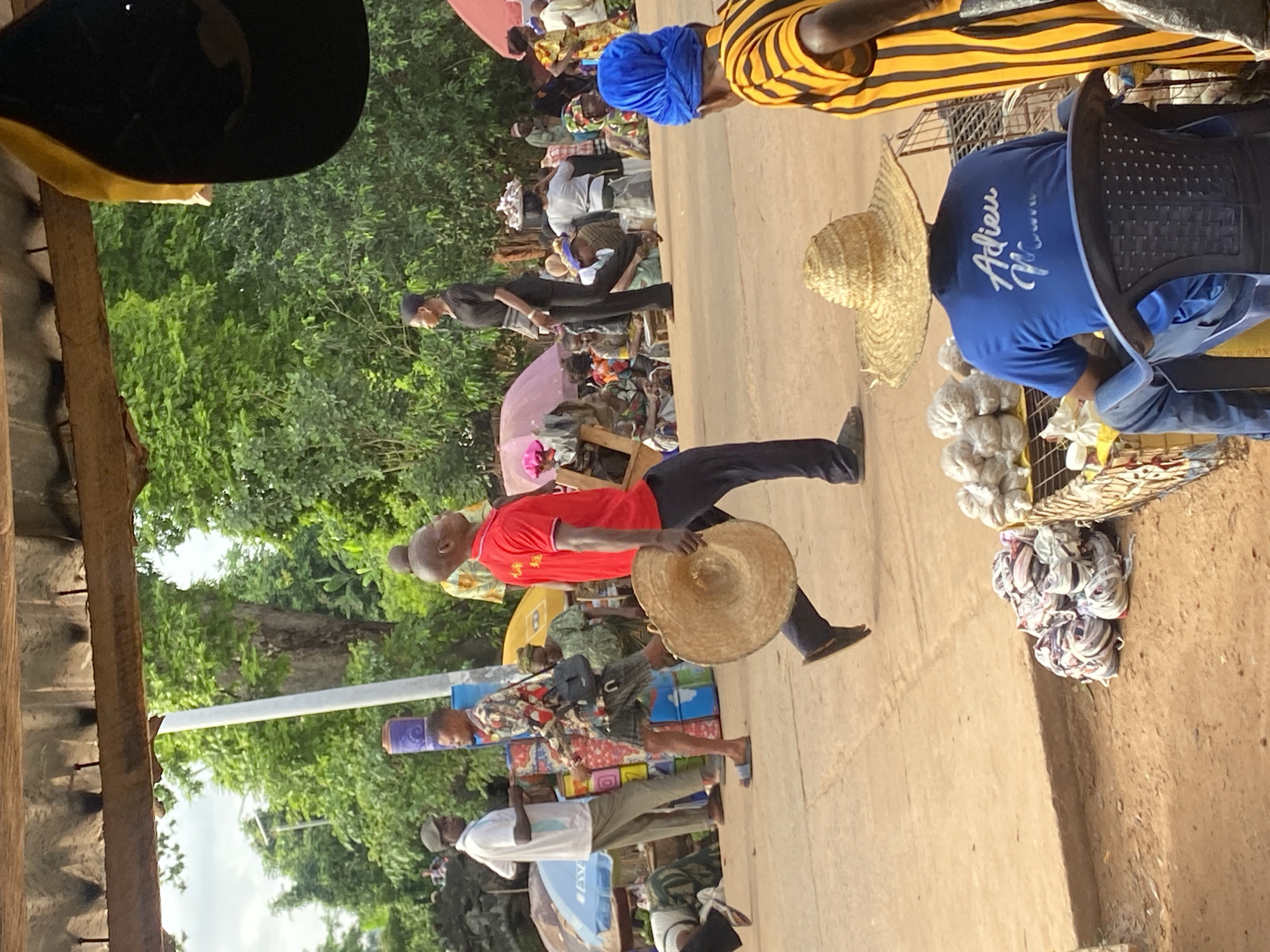 A street scene in Igbara, Nigeria, shows a crowded roadside market. People of varying ages are present. A man wearing a red shirt and dark trousers walks along the edge of the road, carrying a woven sun hat. Another man is wearing a blue shirt with "Adieu" printed on the back. A woman wears a blue head wrap and a yellow striped shirt. Various stalls with products are along the roadside. Trees are in the background.