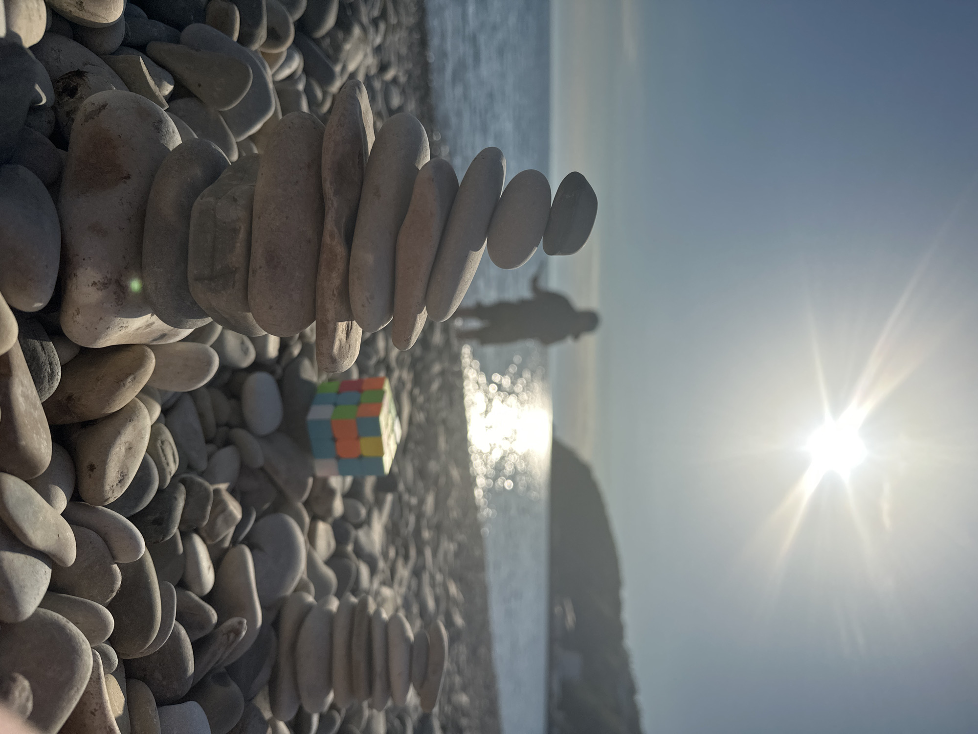 The image captures a stony beach scene, likely in Novomikhaylovsky, Russia, considering the prompt. In the foreground, a stack of flat, oval-shaped stones is balanced on top of other rocks on the beach. Adjacent to the stack, partially visible, is a Rubik's Cube resting on the rocks. In the blurred background, a person's silhouette stands near the water's edge, facing the sea. The sun shines brightly, reflecting on the water's surface. The sky is clear and light blue, suggesting a sunny day. The image is angled, giving a wide perspective of the beach.