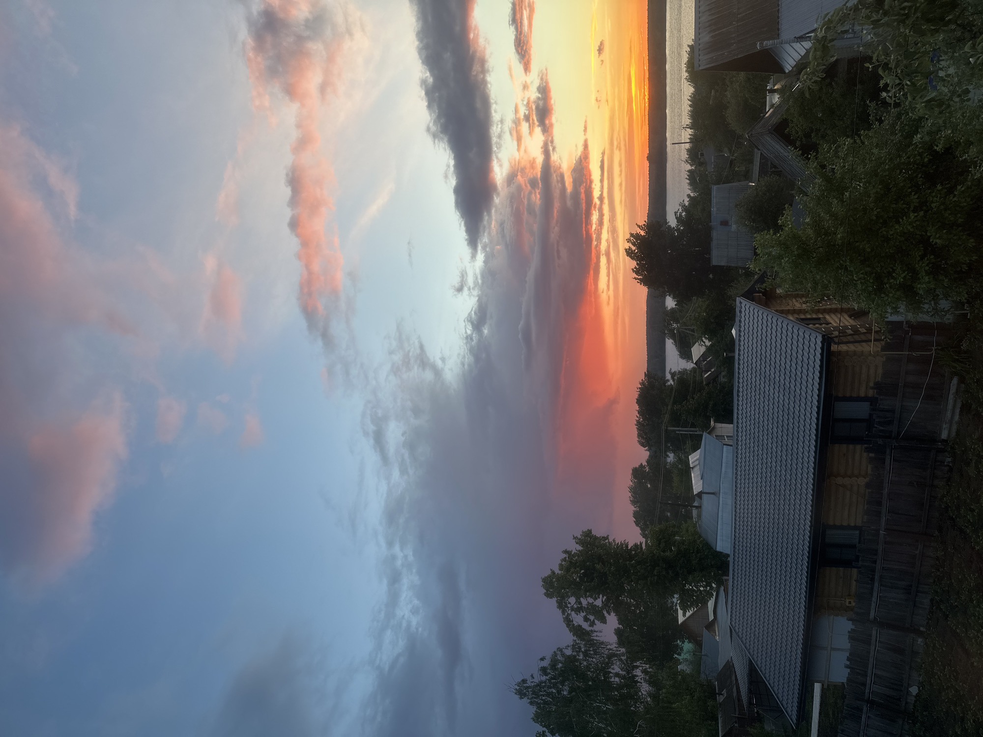 The image captures a sunset in the city of Chaikovsky, Russia. The sky exhibits a gradient of colors, transitioning from blue on the left to orange and yellow near the horizon on the right. Clouds are present, with some displaying pink and gray hues due to the sunset's reflection.
Below the horizon, a body of water is visible, possibly a river or lake. The foreground includes trees and the rooftops of several buildings, indicative of a residential area or village setting. The buildings appear to be constructed from wood, a common architectural style in many parts of Russia. One building has a dark gray tiled roof, suggesting a recent renovation. The image appears to be taken from an elevated position, providing a panoramic view of the landscape.