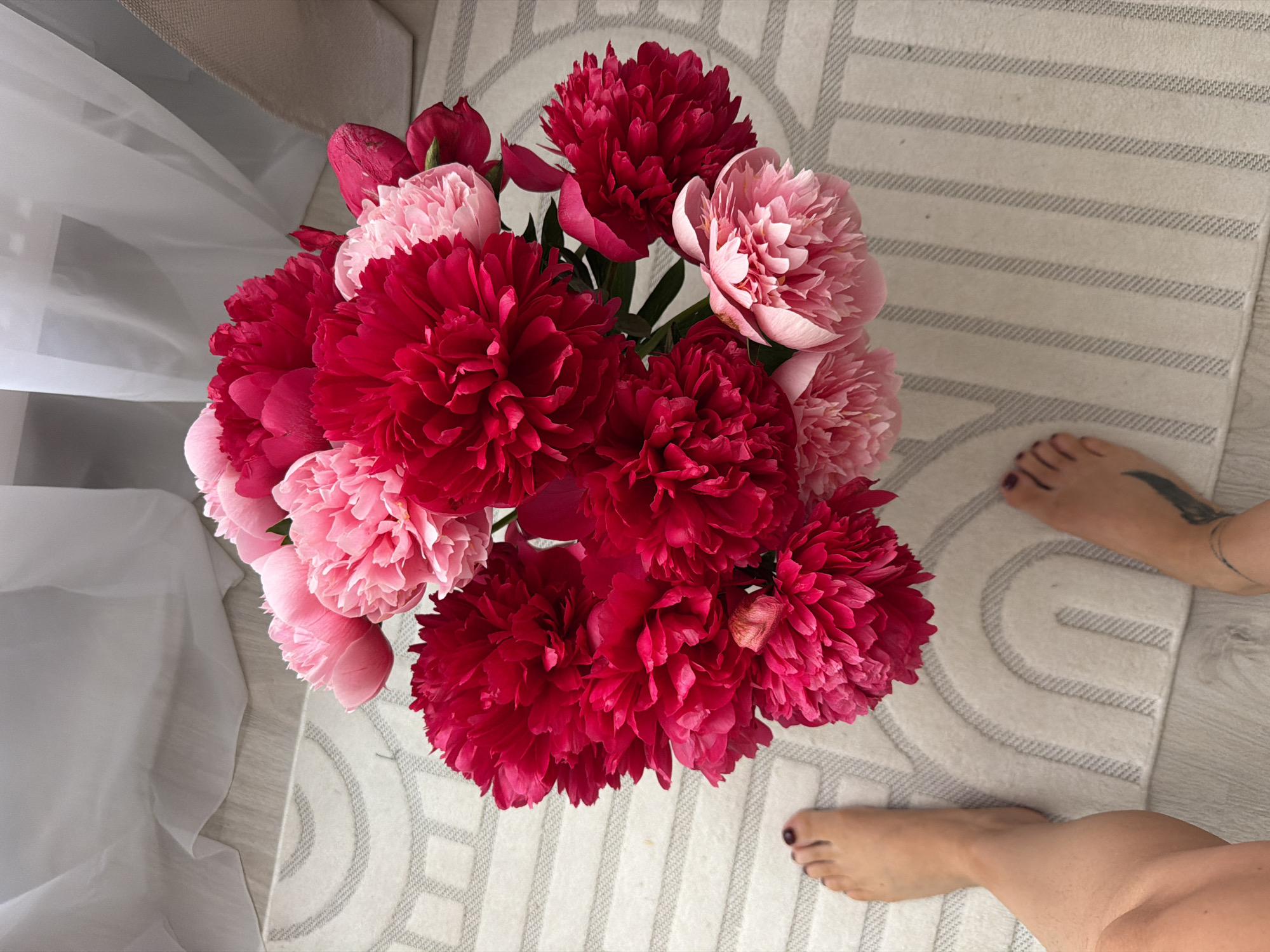 The image shows a close-up, high-angle shot of a bouquet of pink and red peonies on a rug. Two bare feet are visible on the left and right sides of the image, positioned on the rug. The rug has a modern design with arched patterns and stripes. In the background, sheer white curtains are visible, suggesting an indoor setting. The lighting is bright, creating a soft glow on the flowers and the surrounding environment. The scene does not provide any information about the location in Moscow, Russia.