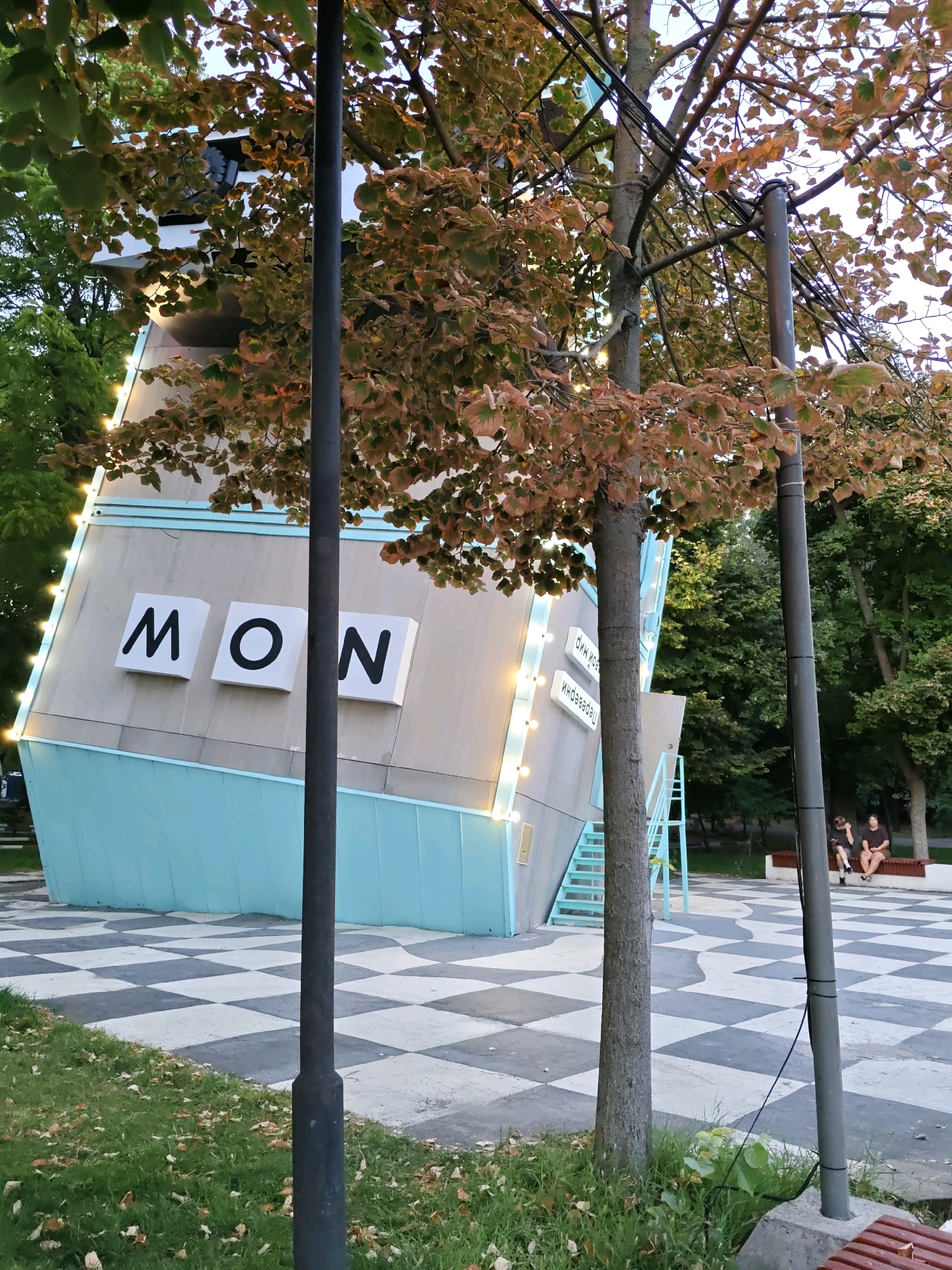 The image shows a modern building with the letters "MON" displayed prominently on its facade. The building is angled, giving it a unique architectural appearance. A patterned ground, resembling a checkerboard with curved lines, surrounds the building. Trees with autumn-colored leaves are scattered around, and there are a couple of streetlights. In the background, two people are sitting on a bench. The scene is set in an outdoor public space, likely a park or plaza. This building is the "MON" (Modern Art Gallery) in Rostov-on-Don, Russia.