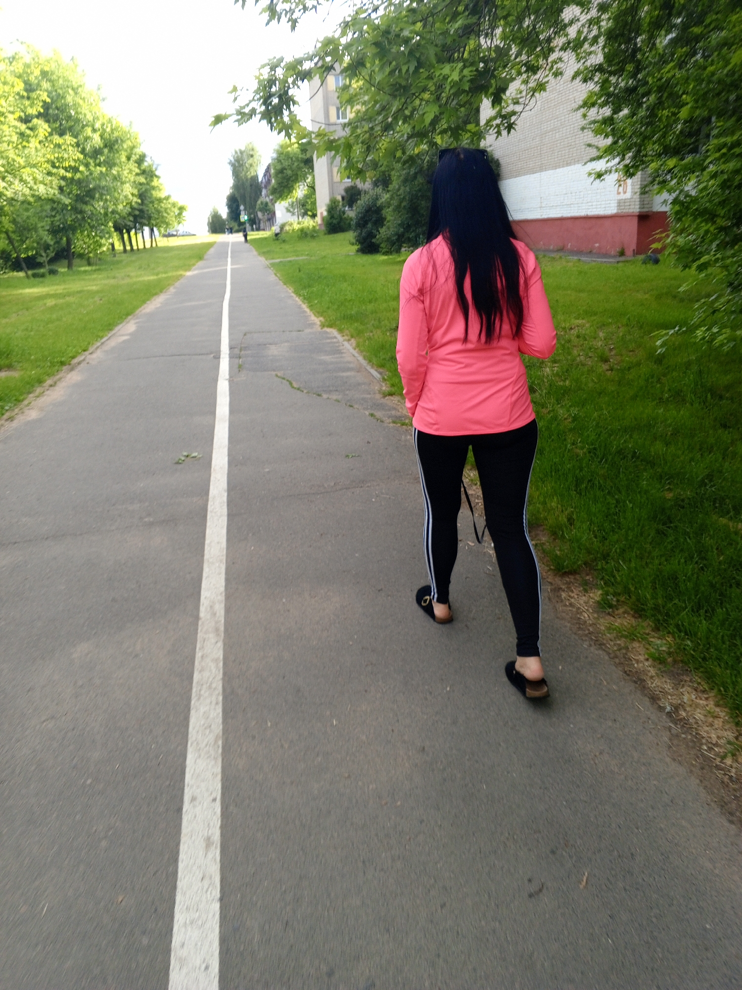 The image shows an outdoor scene in the City of Unknown, Unknown, featuring an asphalt path flanked by grass and trees. A single individual is depicted from behind, walking away from the camera on the path. This person has long, straight, dark hair and is wearing a long-sleeved, coral-pink top, black leggings with two parallel white stripes on the outer seam of each leg, and black open-toed sandals with brown footbeds. A dark strap is visible hanging from their left side.
The asphalt path is gray, exhibiting minor cracks and areas of wear, and is marked by a single, solid white line running down its center. Green grass borders both sides of the path. On the left side, a row of green-leafed trees extends upwards along a gentle incline. On the right side, beyond the grass and trees, a multi-story building is visible. This building features an upper section of light-colored brick and a lower section painted in a distinct red hue. The number "20" and faint additional markings are discernable on the lighter brick section of the building. The sky appears bright and diffused.