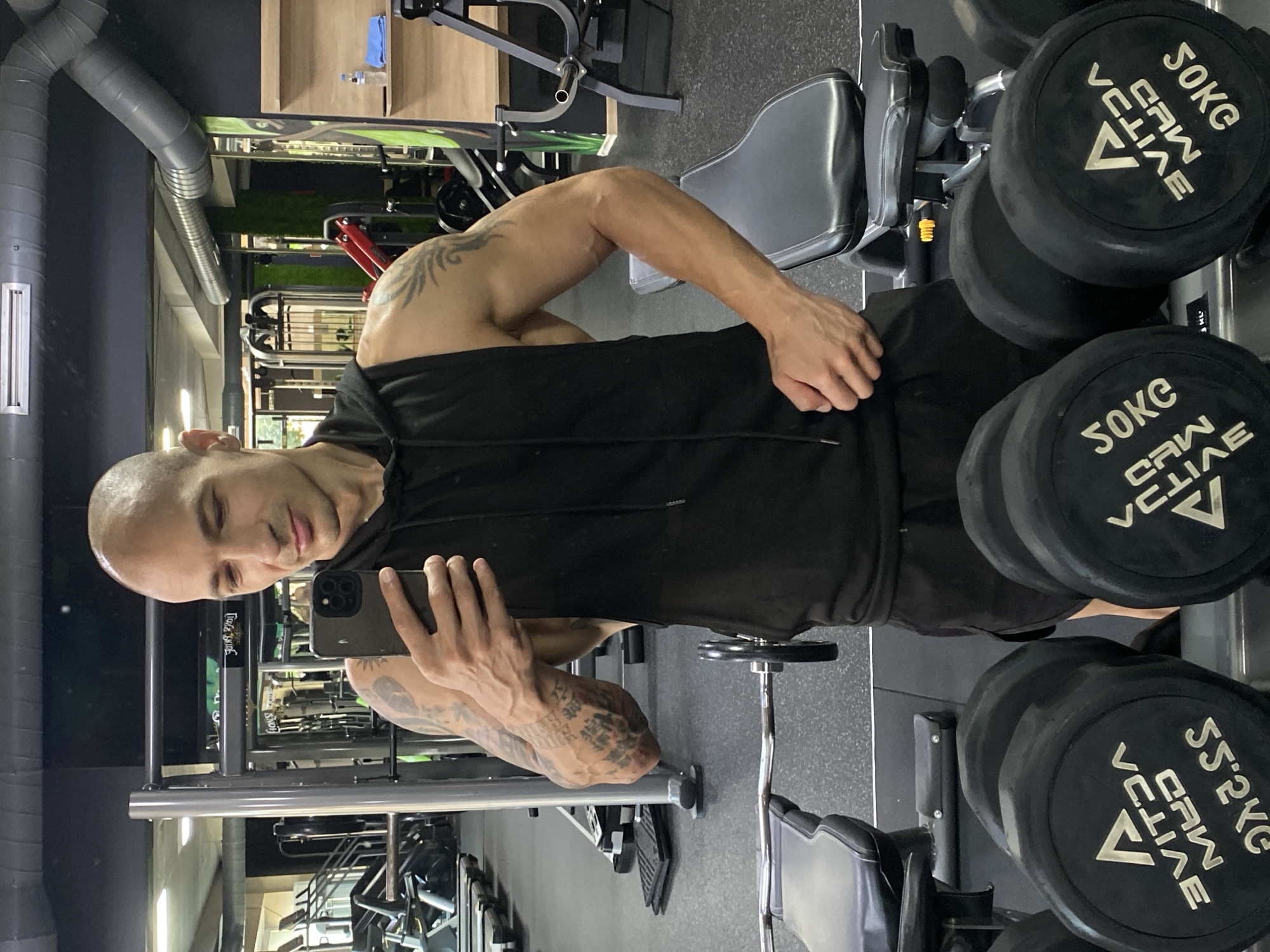 A bald, fair-skinned man with tattoos on his arms is taking a selfie in a gym. He is wearing a black sleeveless hoodie and black shorts. In the background, gym equipment such as treadmills, weight machines, and dumbbells are visible. The gym appears to be located in Pleven, Bulgaria, given the presence of "PLEVEN GYM" signage visible on the wall. A bottle of water and a blue towel are placed on a shelf in the background.