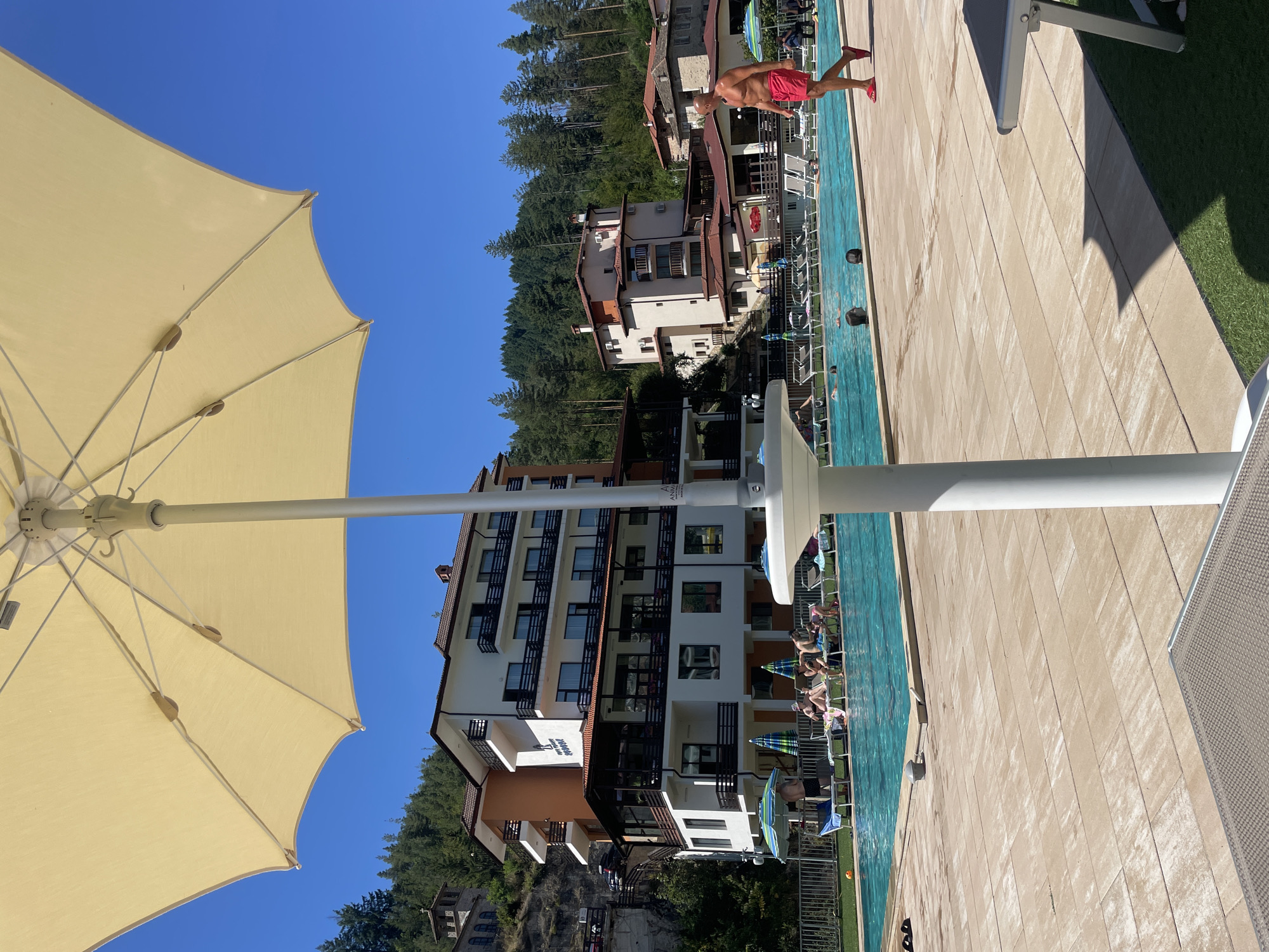 In Banite, Bulgaria, this image captures a poolside scene on a sunny day. A large, light-colored umbrella dominates the foreground, casting a shadow. Beyond it, a group of people relax on lounge chairs near a turquoise swimming pool. Several individuals are in the pool, swimming or wading. On the right side of the frame, a man in red swim shorts walks along the pool deck. In the background, several multi-story buildings with balconies and red-tiled roofs are nestled against a wooded hillside, characteristic of the Rhodope Mountains. The architecture suggests a resort or hotel complex. The sky is clear blue.