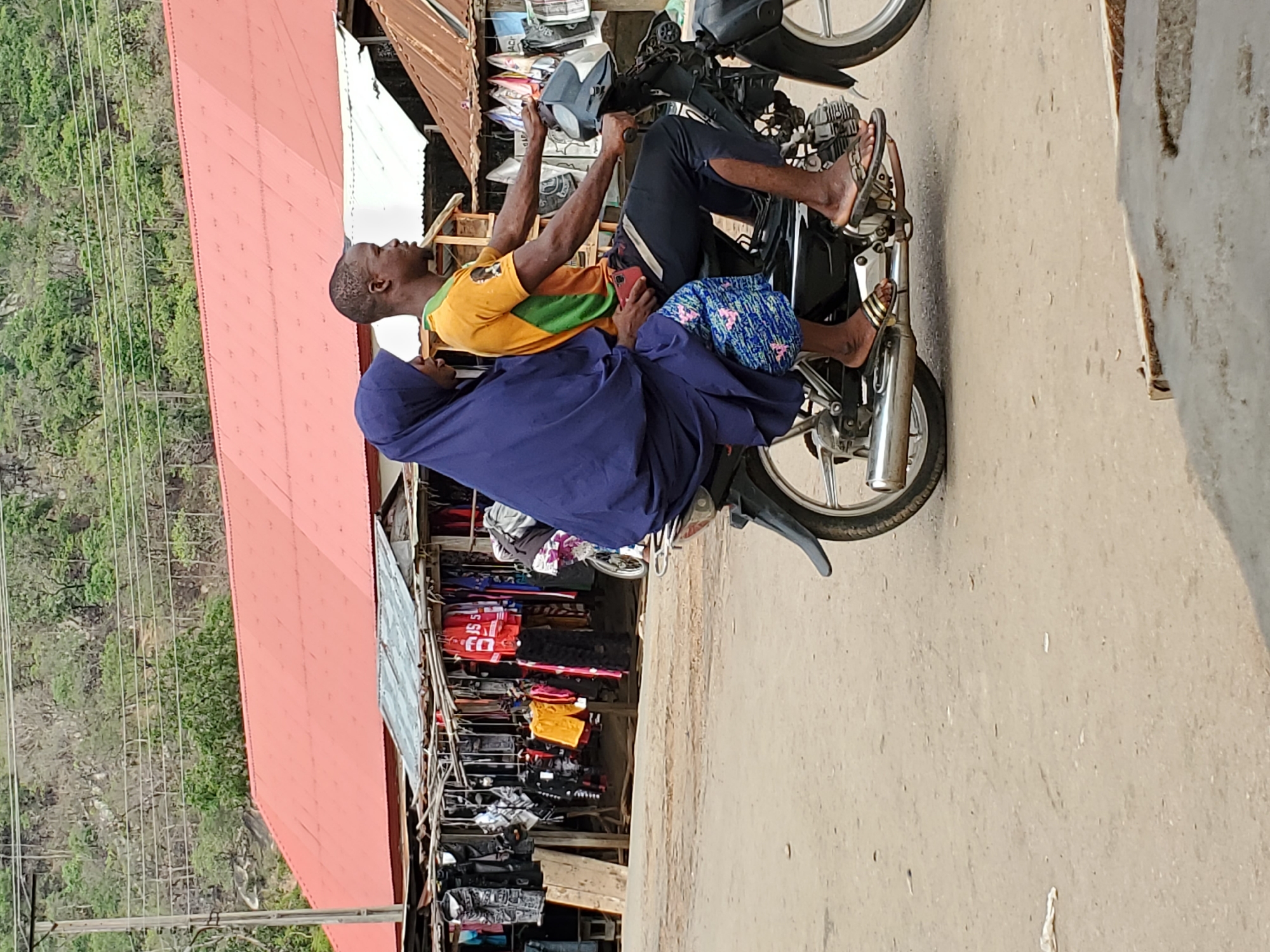 The image captures an outdoor street scene, likely in a bustling commercial area, under bright daylight in Mararaba, Nigeria. Two individuals are seated on a black motorcycle, appearing to be stationary on a light-colored road.
The driver, a dark-skinned man with a short haircut, wears a yellow and green short-sleeved shirt, dark trousers, and sandals. He holds a red smartphone in his right hand, seemingly engrossed in it. Behind him sits a passenger, also dark-skinned, clad in a dark blue garment resembling a hijab that covers their head and body, with colorful patterned fabric draped over their legs. This individual also wears sandals.
The motorcycle features chrome exhaust pipes and spoked wheels. In the background to the left, a lush green hillside or forested area is visible, traversed by utility poles and multiple power lines. To the right, a vibrant market stretches out with numerous makeshift stalls, characterized by corrugated iron roofs. These stalls are packed with various merchandise, including clothing items like t-shirts and shorts, some displaying visible letters and numbers such as 'SS', 'F', and '55' on a red garment. Bags and other goods are also on display.
The clear lighting suggests a sunny day. The overall impression is a common, everyday street scene in a lively urban or semi-urban setting.