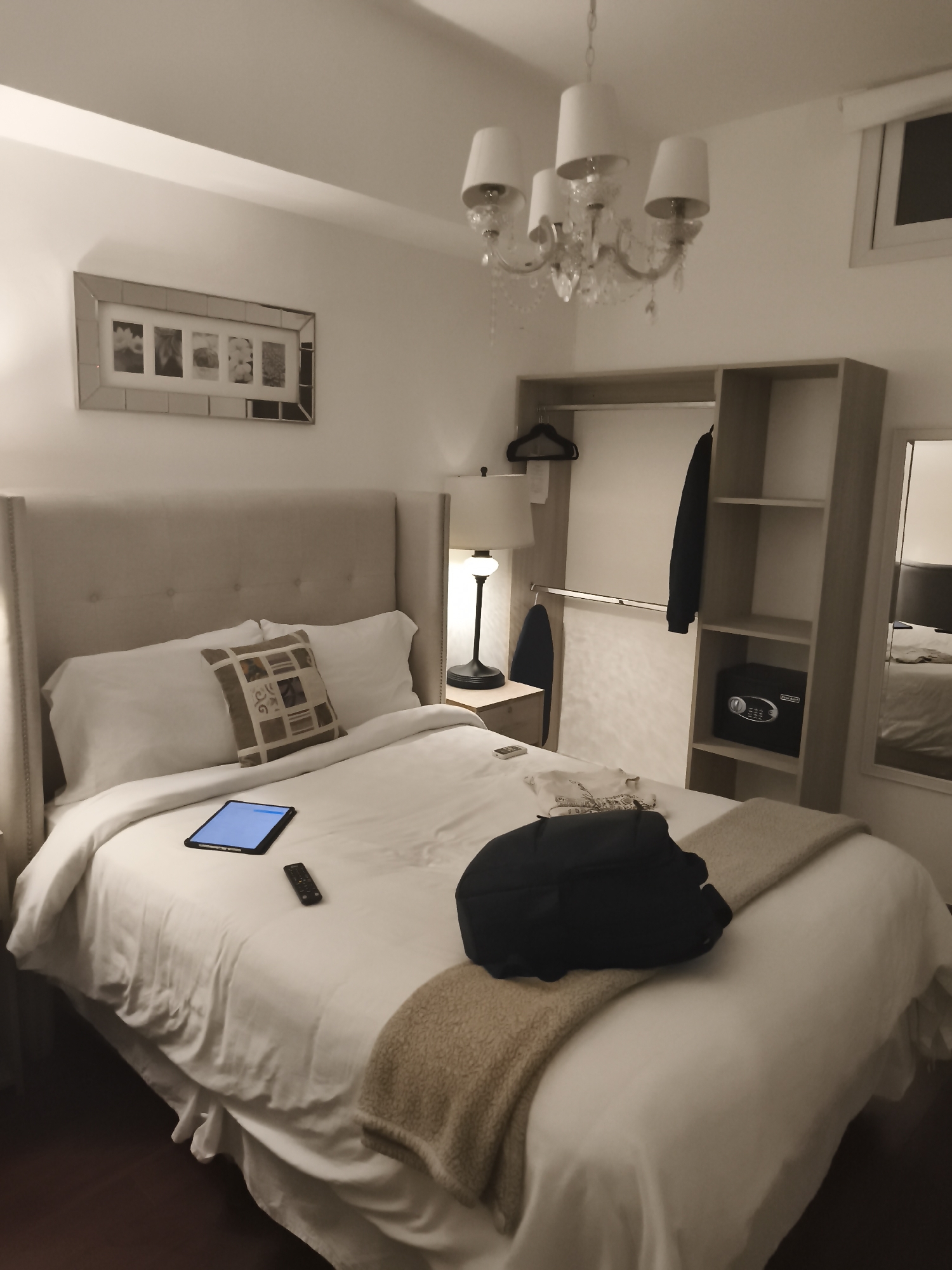 The image shows a hotel room interior. The room features a king-size bed with white linens, decorative pillows, and a light brown throw blanket. A black backpack and remote control are placed on the bed. A blue tablet is also present. A nightstand with a lamp is located next to the bed. A large wardrobe with shelves, a hanging rod, and a safe is against the wall. A chandelier hangs from the ceiling. A picture frame with multiple images is on the wall above the bed. The room has a window, and a mirror is to the right of the wardrobe, reflecting the bed. Guatemala City, Guatemala is the location.