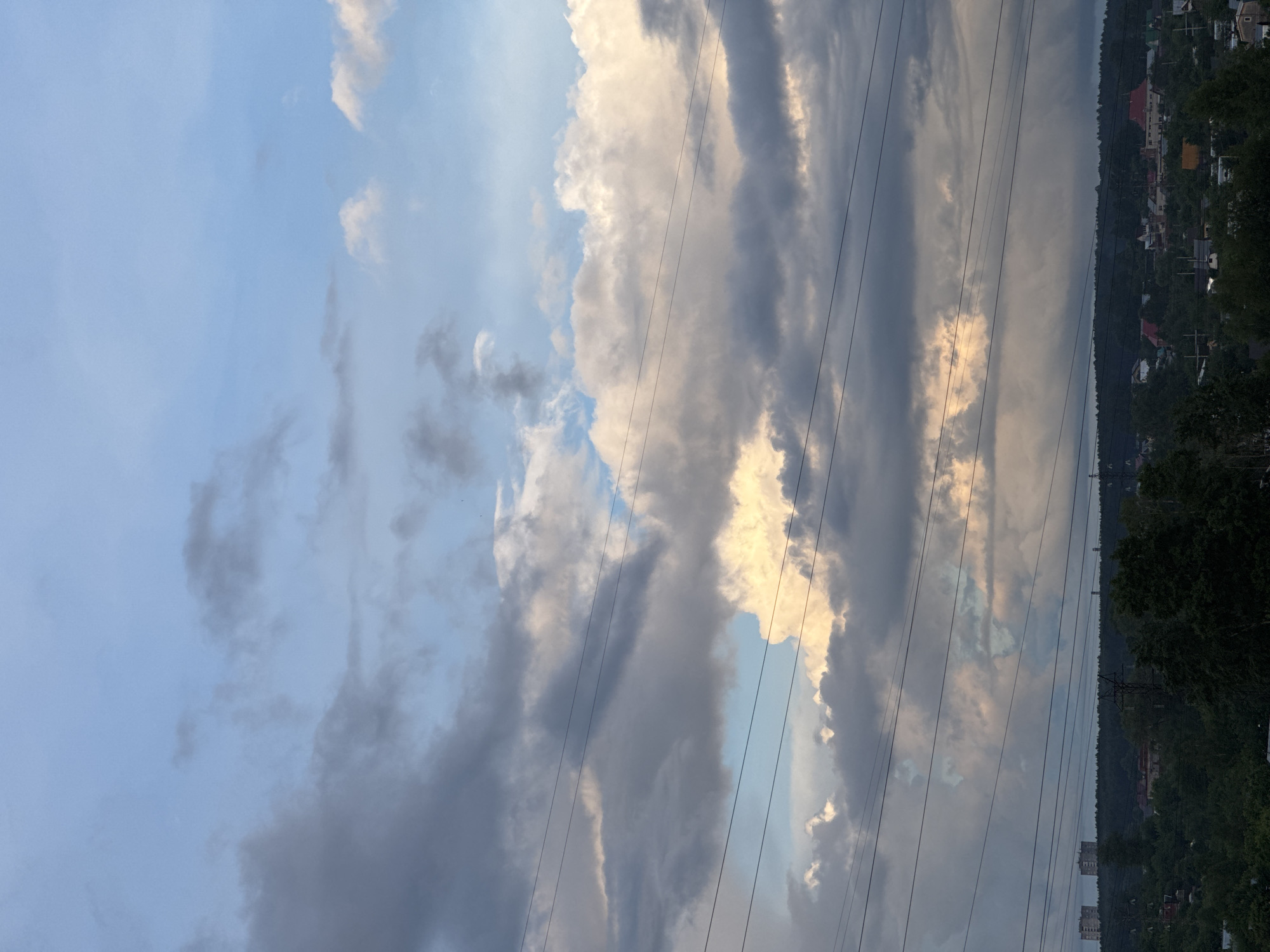 The image captures a vast, dynamic sky dominated by an array of clouds and crisscrossed by power lines, with a distant landscape visible at the bottom. The sky transitions from a light blue hue in the upper left to a dramatic mix of bright, illuminated clouds and dark, heavy grey formations across the rest of the frame. Some clouds glow with a warm, golden-white light, likely from a low-angled sun, suggesting late afternoon or early evening. These brighter clouds contrast sharply with the dark, dense clouds that imply substantial cloud cover, possibly hinting at impending weather.
Several thin, dark power lines stretch horizontally across the mid-ground and upper sections of the image, running parallel to each other and cutting across the cloudy sky.
Below the sky and power lines, a distant landscape unfolds. A dense line of dark green trees forms a forested area, likely a hillside or riverbank, stretching across the horizon. Amidst these trees and on the distant slopes, numerous small houses and buildings are visible, some identifiable by their distinct red roofs. Towards the far left of the landscape section, larger, possibly multi-story buildings can be discerned.
There are no people or explicit activities taking place. No visible text is present. The overall scene depicts a natural landscape with human infrastructure, all under a captivating and dramatic cloudy sky in Chaikovsky, Russia.