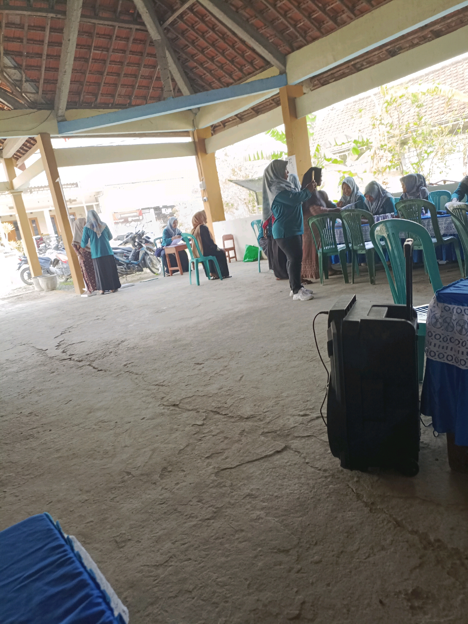 The image depicts an outdoor gathering under a covered structure, likely in Rancang Kencono, Indonesia. Several individuals are seated at tables, some wearing headscarves and engaged in conversation or activity. Others are standing, with one individual in a teal top and dark pants actively using a smartphone. In the background, motorcycles are parked, suggesting a local community event or meeting. The environment is characterized by a concrete floor, wooden support beams, and a tiled roof overhead. A portable speaker is positioned in the foreground, and tables covered with blue tablecloths are visible. The overall scene suggests a casual, informal gathering.