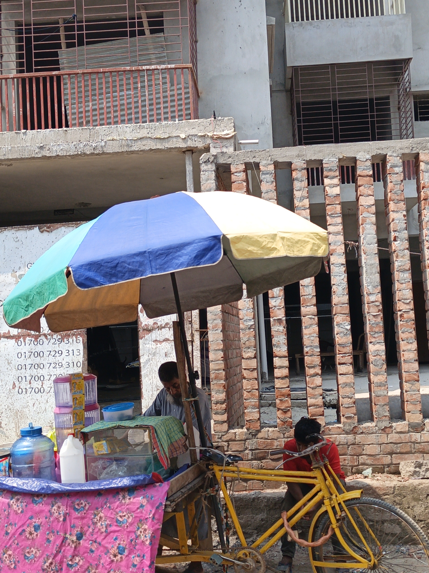 A man in a gray shirt stands behind a mobile vendor cart, operating it under a multicolored umbrella. The cart, draped with a floral pink cloth, displays several containers, including water jugs and plastic dispensers. A yellow bicycle is connected to the cart. Another person, wearing a red shirt, is seated next to the bicycle. The background features a partly constructed brick building with a series of vertical brick columns. A structure with a red railing is visible in the upper left. Phone numbers are painted on a wall near the cart. The location appears to be Dhaka, Bangladesh.