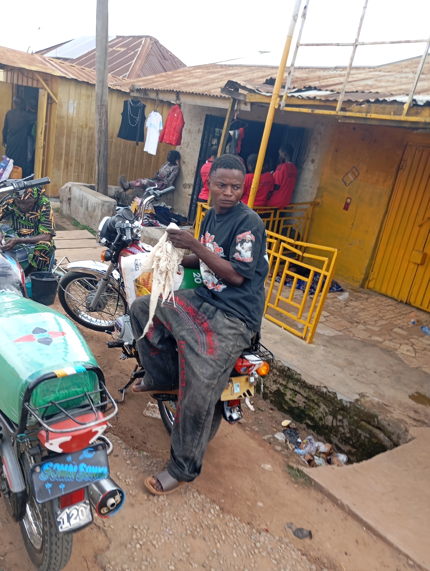 In Shendam, Nigeria, a young Black male sits on a motorcycle, holding a white cloth. He wears a black t-shirt with graphics and distressed grey pants. He is positioned in front of a row of small shops with corrugated metal roofs. Behind him, various items are displayed, including clothing items hanging from a line. Another motorcycle with a green and white cargo box is partially visible to the left. The ground is dirt and gravel. In the background, people can be seen inside the shops. A yellow metal barrier is present in front of one of the shops. A ditch filled with debris is located to the right of the motorcycle the man is sitting on.