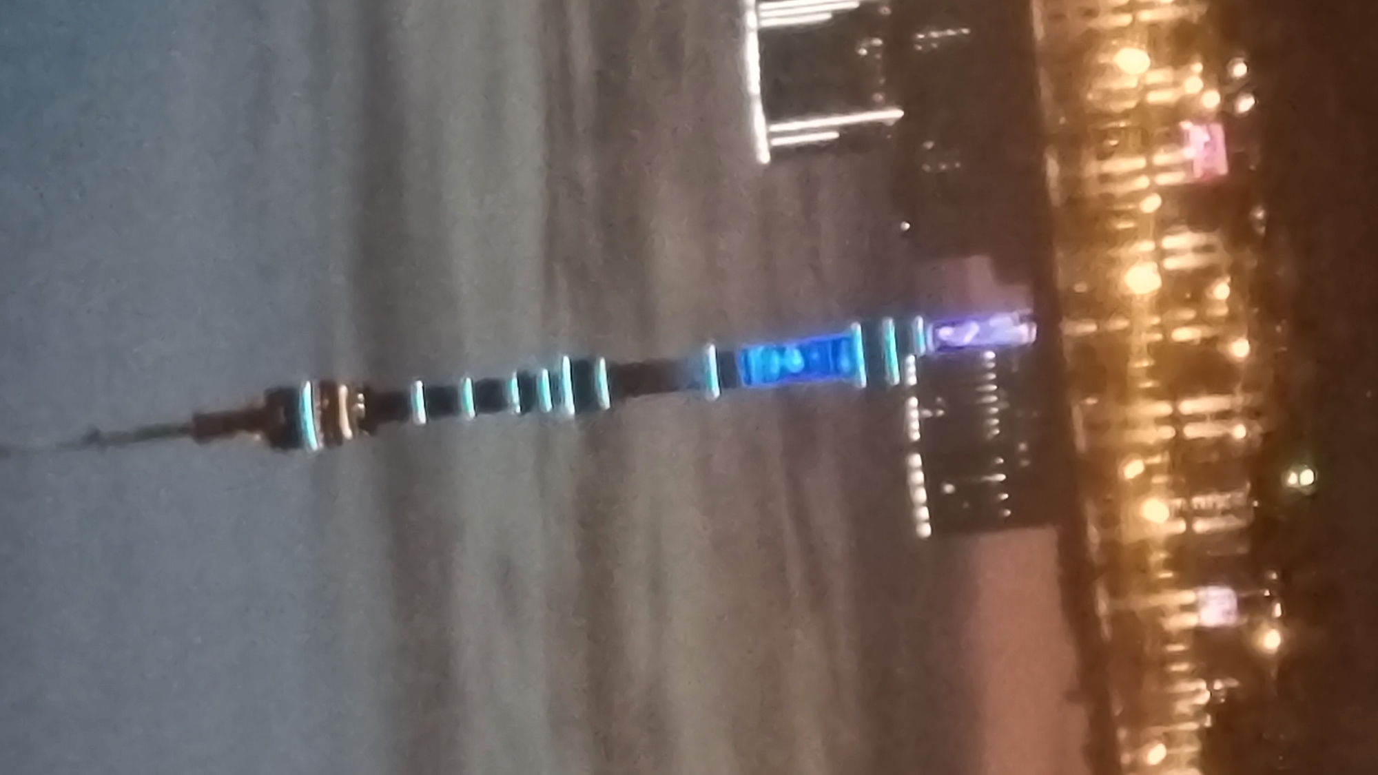 The image shows the Ostankino Tower in Moscow, Russia at night. The tower is illuminated with blue and white lights. In the foreground, there is a cityscape with many buildings and lights. The sky is dark and cloudy, with some hints of pink and orange from a sunset or twilight. The image is slightly blurry, suggesting it was taken from a distance or with a low-quality camera.