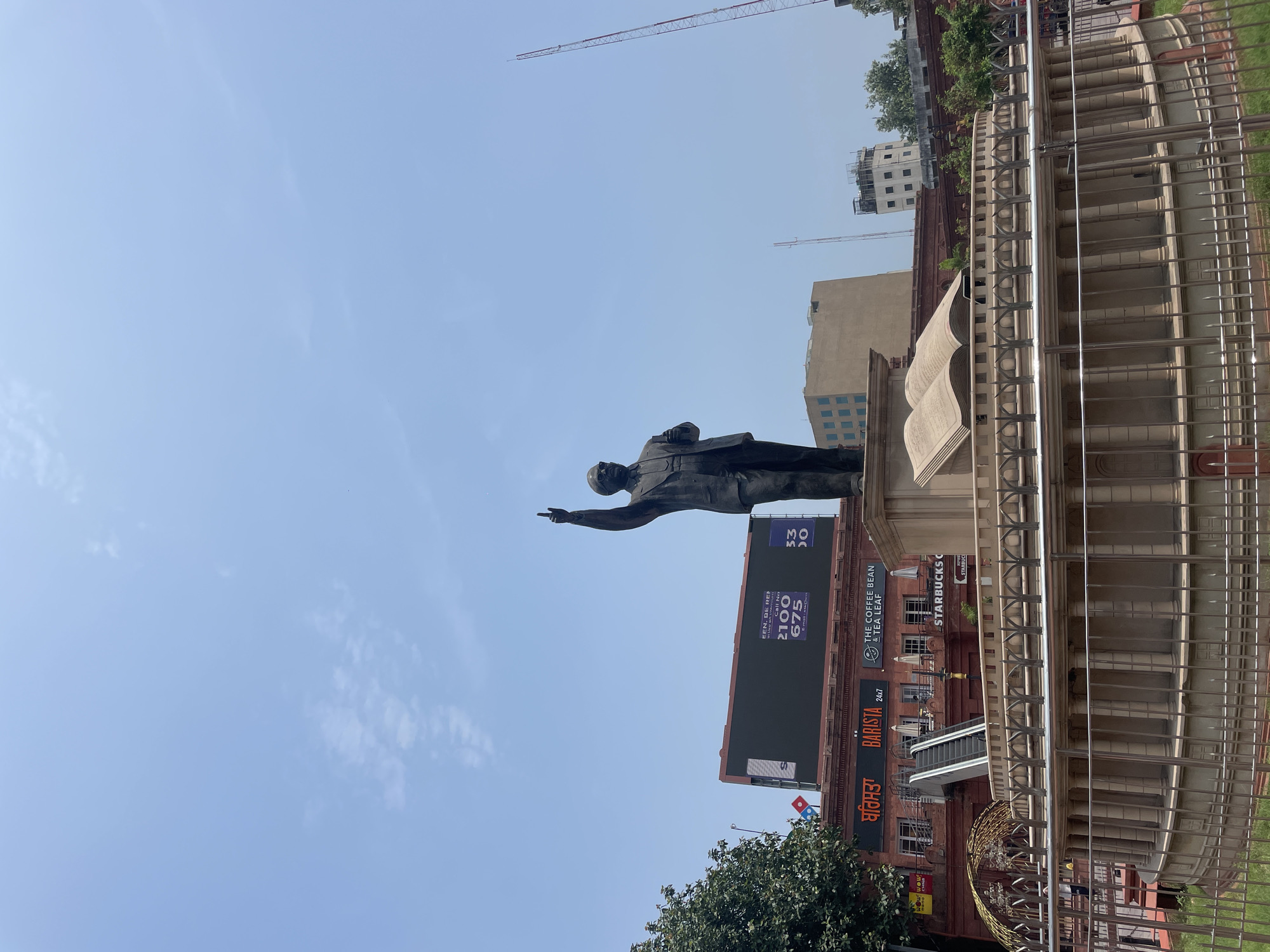 The image shows a statue of B.R. Ambedkar, a prominent figure in Indian history, located in Amritsar, India. The statue stands on a pedestal which includes an open book, symbolizing knowledge and education. Behind the statue, a large brick building houses several businesses, including "Barista" and "The Coffee Bean & Tea Leaf," as well as a Starbucks. A large digital billboard is also visible on the building, displaying numbers and text. The scene is set against a clear blue sky with a few wispy clouds. In the background, several tall buildings and communication towers can be seen, indicating an urban environment. The foreground features a metal railing and lush green grass. The overall impression is that of a public monument in a bustling city.