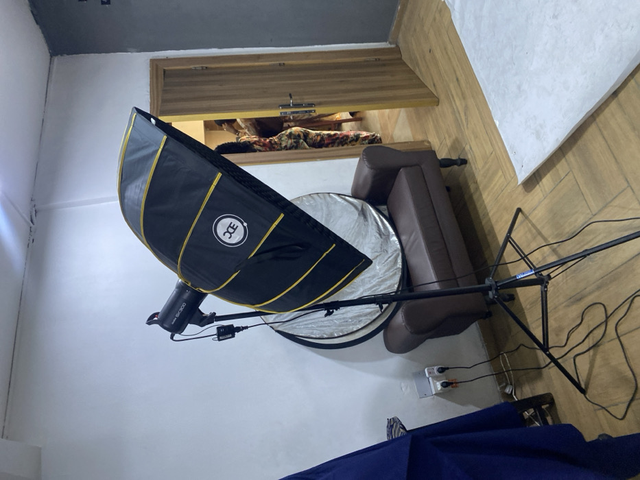This media file presents an indoor scene, rotated 90 degrees to the left, appearing to be a temporary photography setup in Animashawun, Nigeria.
The primary subjects are photography equipment arranged in a room. In the foreground, dominating the left side of the frame, is a large black photography softbox with prominent gold stripes and a white circular "DC" logo. Attached to it is a strobe light, partially visible, with "SK300" and likely "GODOX" text on its body. Below and slightly behind the softbox, a circular silver photography reflector stands on the floor.
To the right of the softbox and reflector, a brown leather two-seater sofa is visible. Extending from the bottom right towards the center of the image is a black light stand or boom arm, with various power cords trailing from it to an electrical outlet mounted on the light-colored wall.
In the background, there's a wooden structure, possibly a door or a large cabinet, positioned above the sofa. Behind this wooden element, a pile of patterned textiles or clothing (brown and floral) is discernible. The floor is covered with light-colored wooden planks, and the walls are white or off-white, with a darker ceiling visible in the upper left corner. The setup suggests that a photography session is either being prepared, is in progress, or has recently concluded in this space. No people are visible in the image.