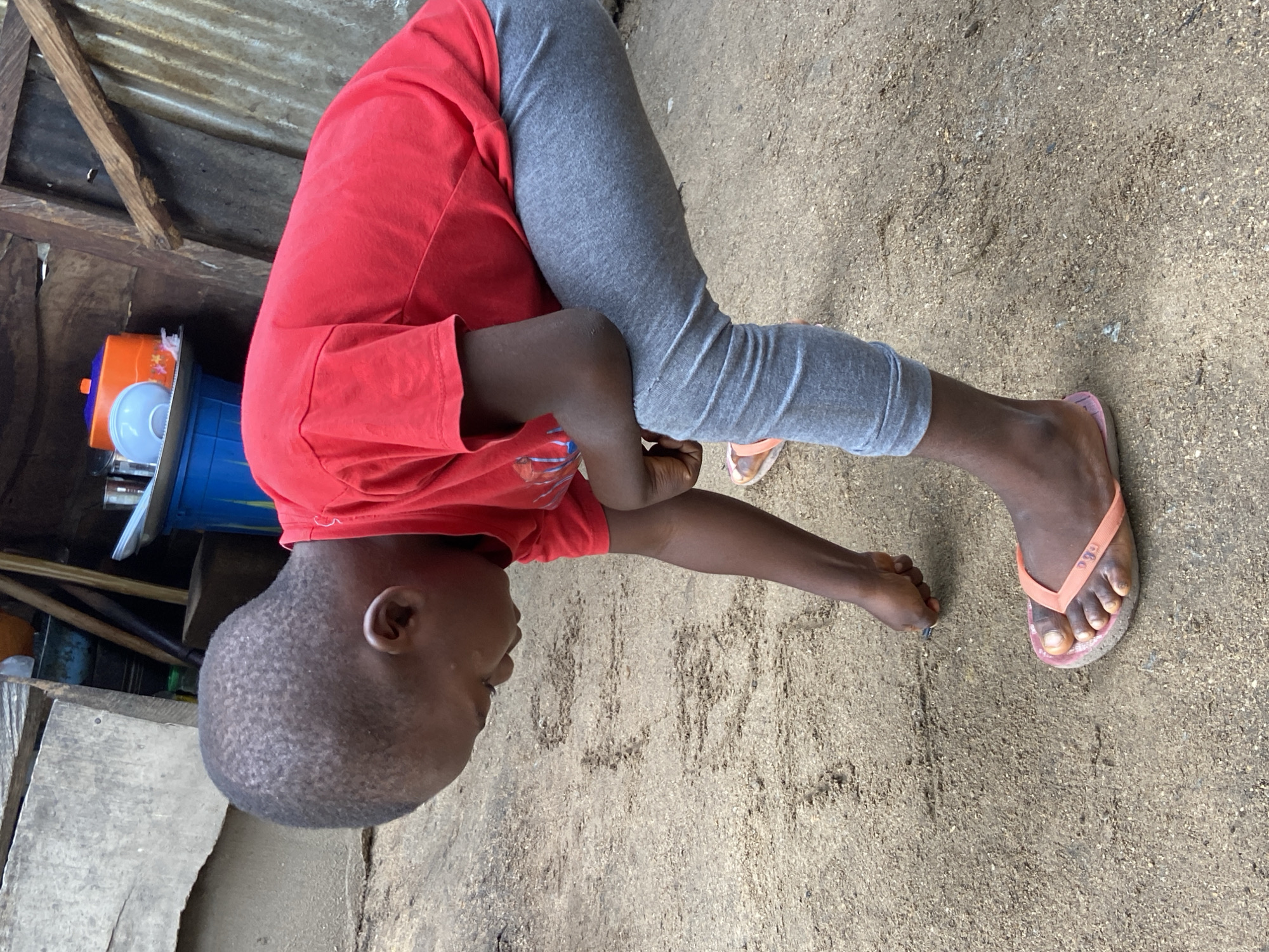 In Mararaba, Nigeria, a young child with dark skin and closely cropped hair is depicted outdoors, bent over and focused on the ground. The child wears a red short-sleeved t-shirt, grey leggings, and orange flip-flops. With their right hand, they are using a small, dark, pen-like object to draw or write letters in the dry, light brown sandy soil; visible markings include partial letters "O", "M", and "N". The immediate background features rudimentary structures: a large, light grey concrete slab and wooden planks to the left. To the right, domestic items are stacked, including a blue plastic bucket, a silver tray, an orange lidded container, and a light blue bowl. Behind these objects, a corrugated metal sheet serves as a wall, with a wooden beam extending diagonally above it. The scene is illuminated by ambient daylight.