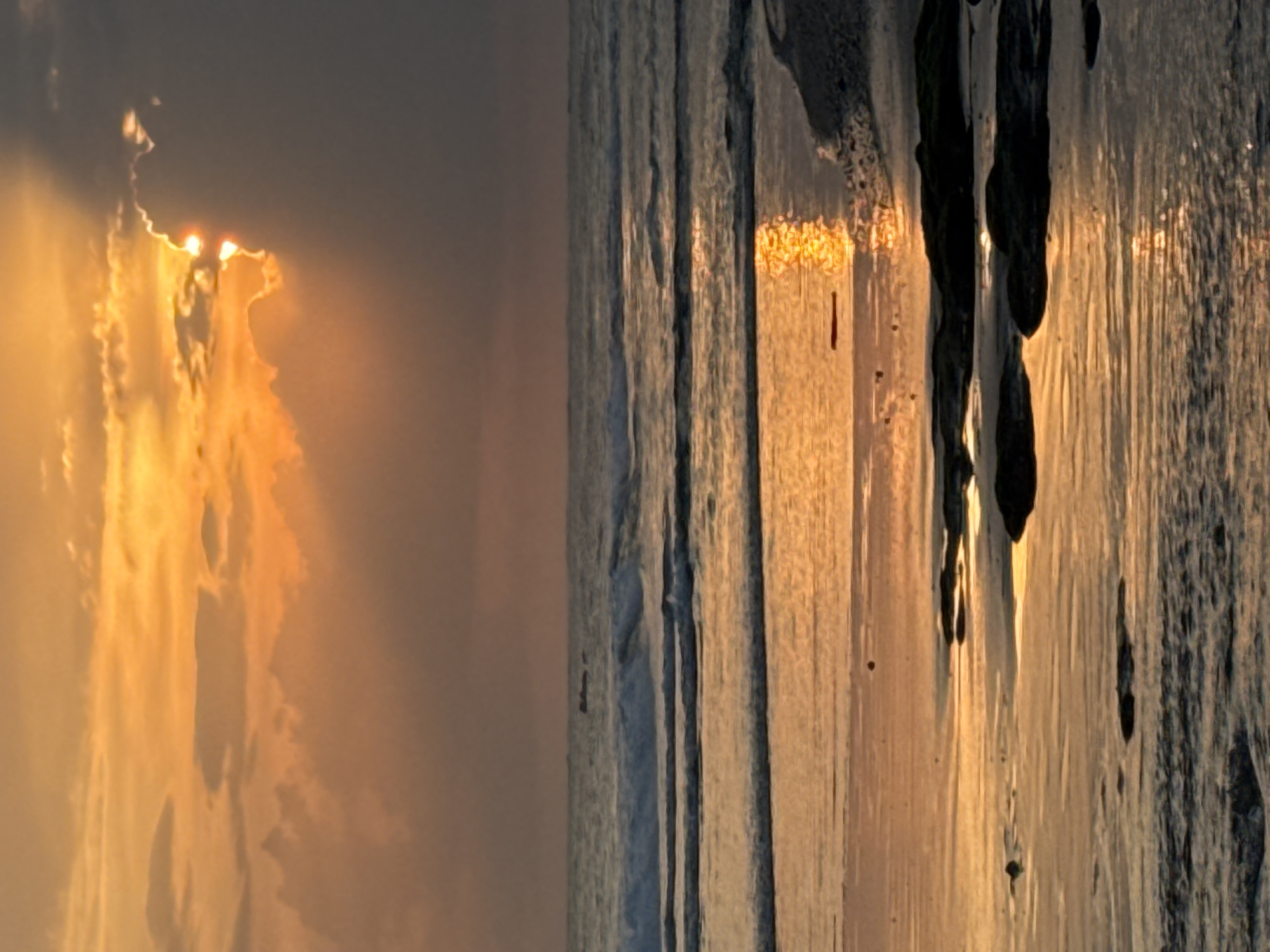 This is a photograph of a sunset over the ocean, with a view of the sky and the water. The sun is partially obscured by clouds, casting a warm, golden light on the scene. The clouds are illuminated from behind, creating a dramatic effect. The water is calm, and the reflection of the sun can be seen on its surface. There are dark, vertical shapes in the foreground, which could be driftwood or other debris on the beach. The overall mood of the image is peaceful and serene, with a sense of awe at the beauty of nature. The time of day is sunset, and the weather appears to be clear, with some clouds. The location is likely a beach in Mengwi, Indonesia, given the tropical setting and the presence of the ocean. There is no visible text in the image.