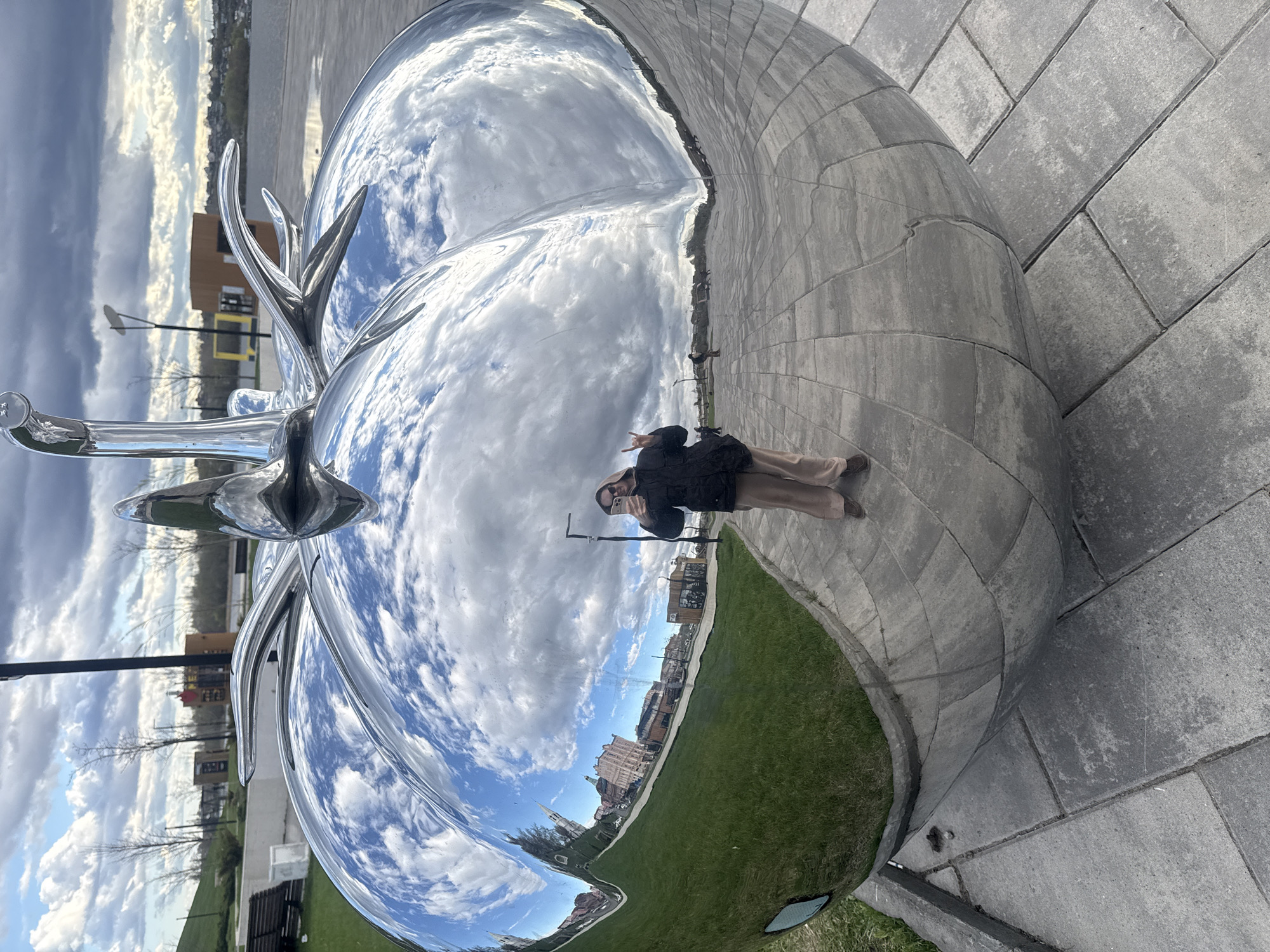 A woman stands holding a smartphone in front of a large, reflective, silver sculpture resembling an apple. The sculpture reflects the sky, clouds, and surrounding cityscape. The woman is wearing a dark jacket and light-colored pants. The ground around the sculpture is paved with light gray concrete blocks. Green grass is visible along the edge of the pavement. The sculpture is located in Syzran, Russia.