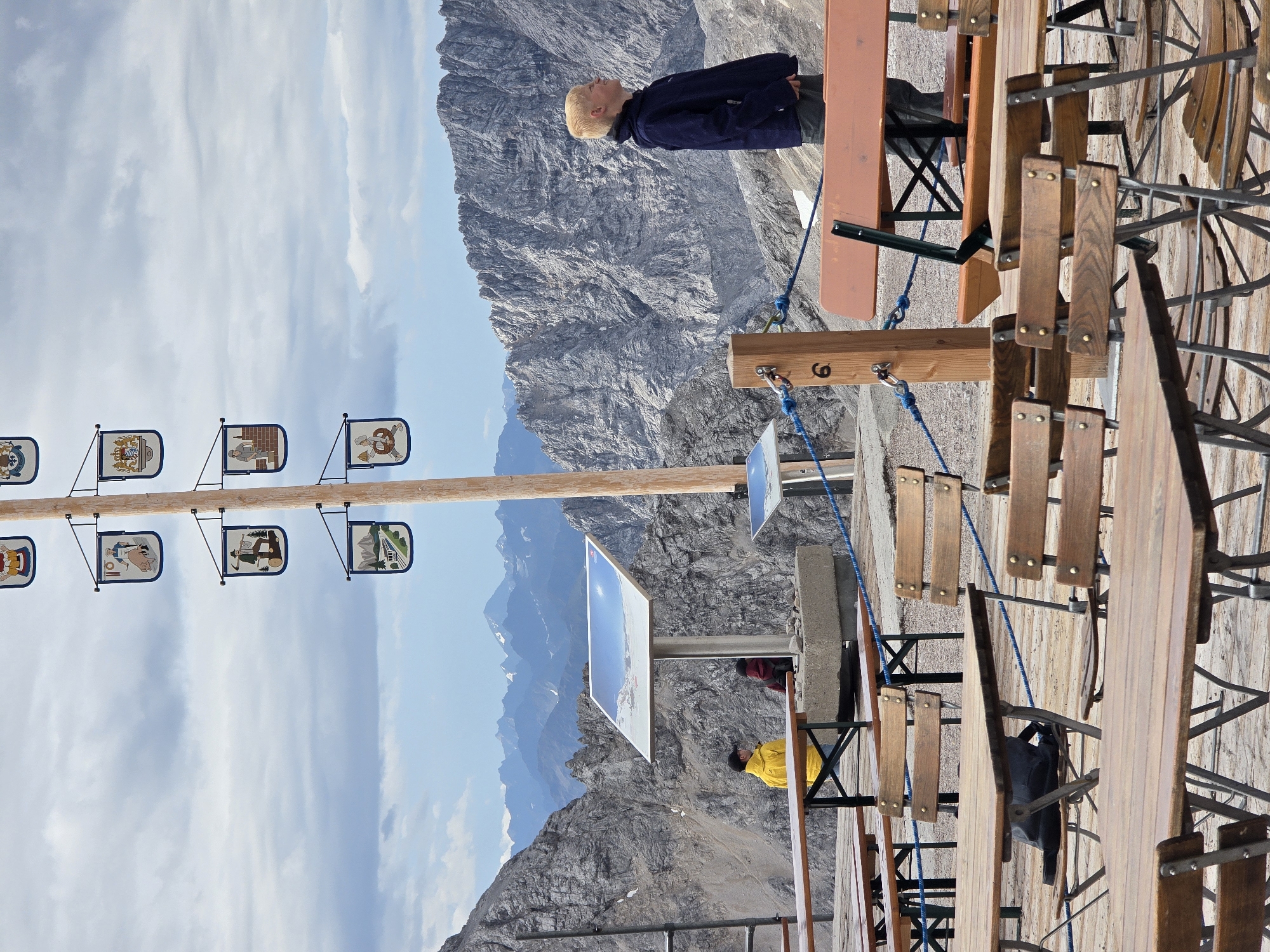 The image depicts an outdoor seating area with wooden tables and benches set against a mountainous backdrop. A tall wooden pole adorned with several small, decorative flags is prominent in the center of the frame. A young boy with blonde hair, wearing a dark jacket and pants, stands facing to the right, looking out at the scenery. In the background, a person in a bright yellow jacket is visible near a information board. The mountainous terrain suggests a high-altitude location, likely in the Alps, consistent with Garmisch-Partenkirchen, Germany, known for its mountainous landscape and tourist attractions. The sky is overcast with clouds.