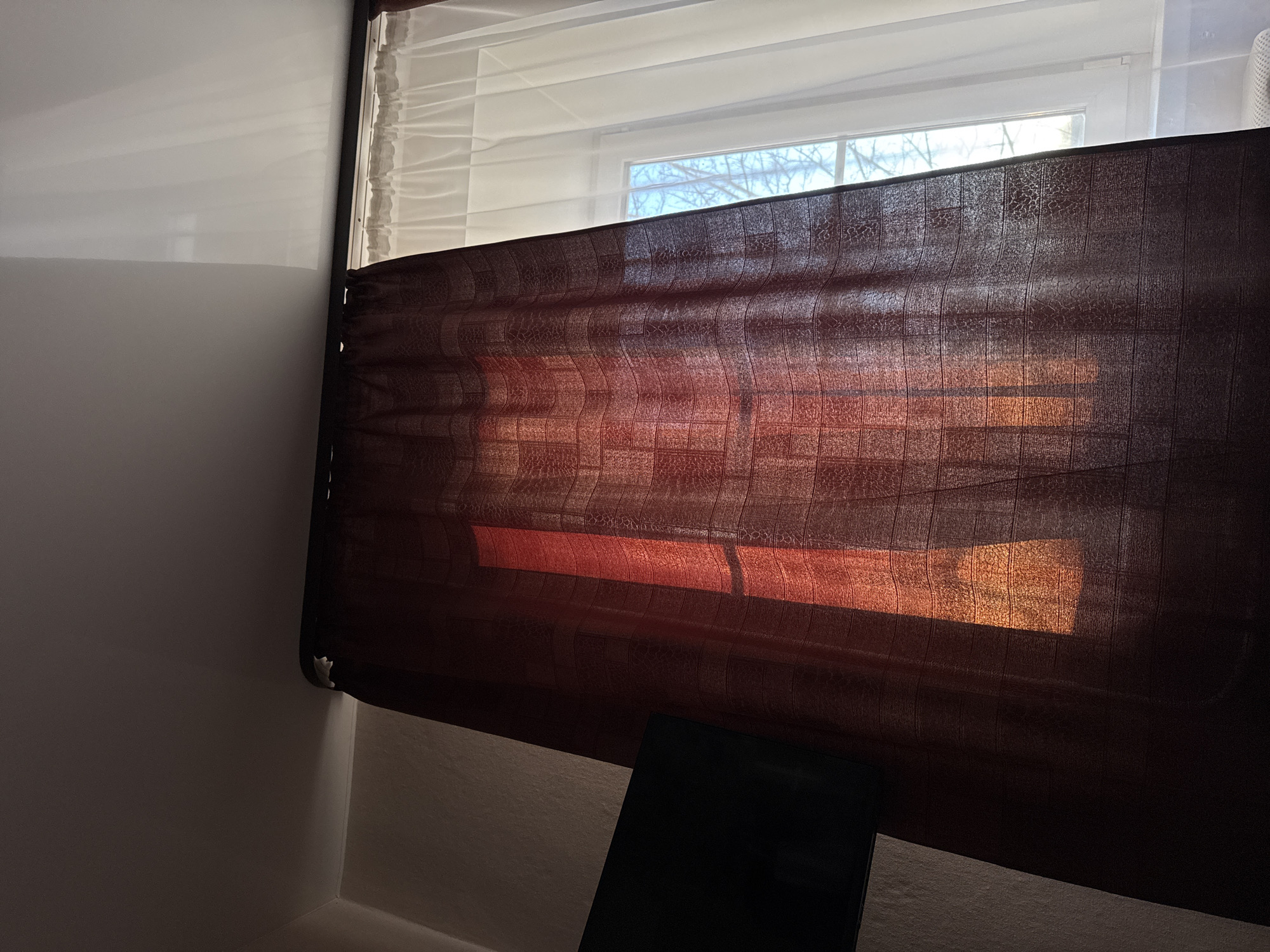 A partially drawn, brown fabric window shade covers a window in an indoor space. A sheer white curtain is visible behind the shade. The window reveals a wintery scene with bare tree branches. The light filtering through the shade casts patterns on its surface. In the foreground, a black rectangular object, potentially a laptop or screen, is partially visible. The location appears to be indoors, potentially in a home or office. The given city Chaikovsky, Russia, is likely irrelevant to the details visible in the image.