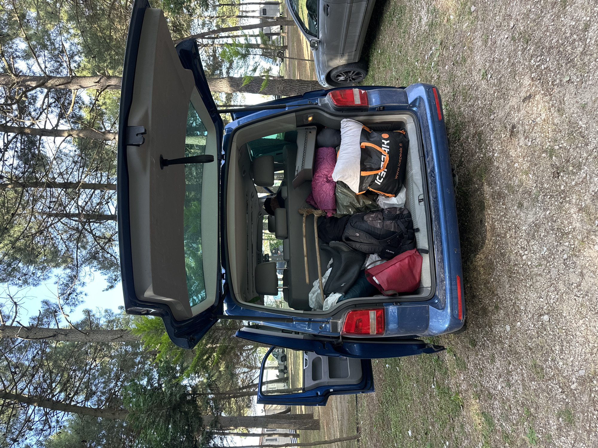The image shows the open trunk of a blue Volkswagen Transporter, heavily packed with luggage and camping gear. An "Icepeak" branded bag is visible among the items. The rear passenger-side sliding door is also open. The car is parked in a wooded area with a gravelly surface, likely a campground. A silver car is parked behind the blue van. The location is Donji Štoj, Montenegro, indicated by the GPS coordinates. The environment appears to be sunny, with tall pine trees visible in the background. No people are visible in the image.