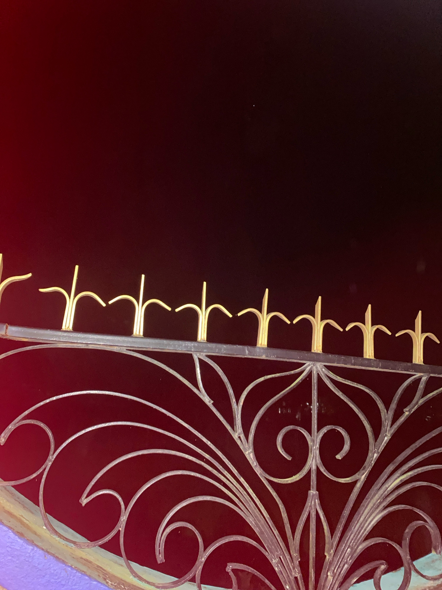 The image shows a close-up of ornate metalwork, likely a gate or fence, set against a deep red background. The metalwork is a combination of dark, possibly black, metal for the main structure and accents of gold for the decorative elements at the top and within the scrollwork. The top edge of the metalwork features a row of pointed, gold-colored finials. Below this, a detailed pattern of silver-colored scrollwork, reminiscent of Art Nouveau or Victorian design, is visible. The scrollwork includes swirling lines, leaf-like shapes, and a central heart-shaped motif. The background is a dark, rich red, which transitions to black in the upper portion of the image, creating a sense of depth and drama. The lower edge of the image reveals a sliver of what appears to be a purple surface, possibly a wall or another part of the structure, painted with some textured or chipped detailing. The scene appears to be an exterior shot, possibly at dusk or in low light, given the dark background and the way the light catches the metal. The location of Naksalbari, India, is not visually represented in the image; it is an external piece of information provided.