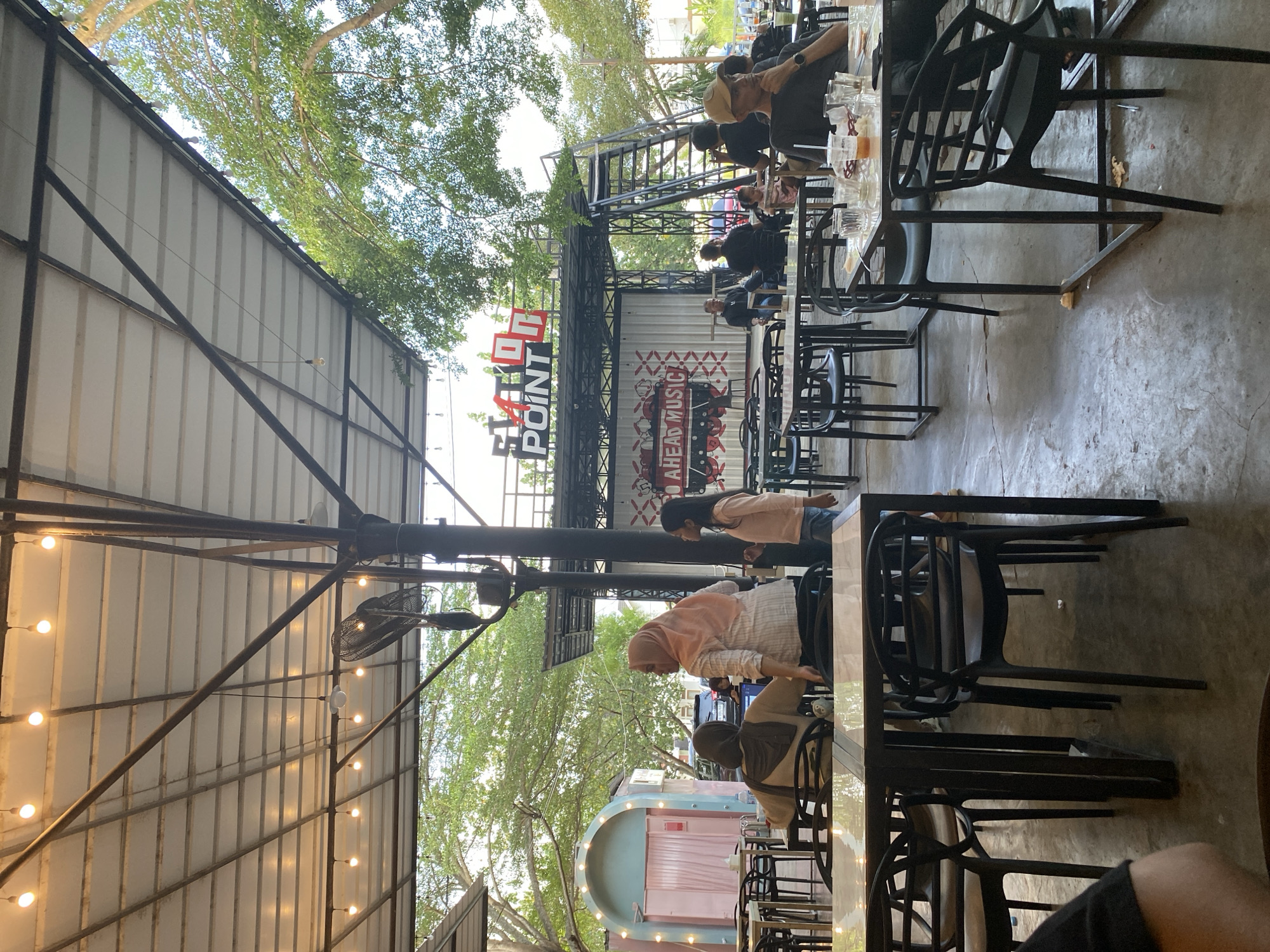 The image depicts an outdoor dining area under a metal roof structure adorned with string lights, likely in Lhokseumawe, Indonesia. Black metal chairs and tables are arranged throughout the space, occupied by several individuals. Near the center of the frame, two women, one wearing a hijab, stand at a table. Further back, towards the center of the scene, a multi-level structure with a sign reading "FATON POINT" is visible. People are seated at tables in the background as well. Trees surround the dining area. The floor is concrete.