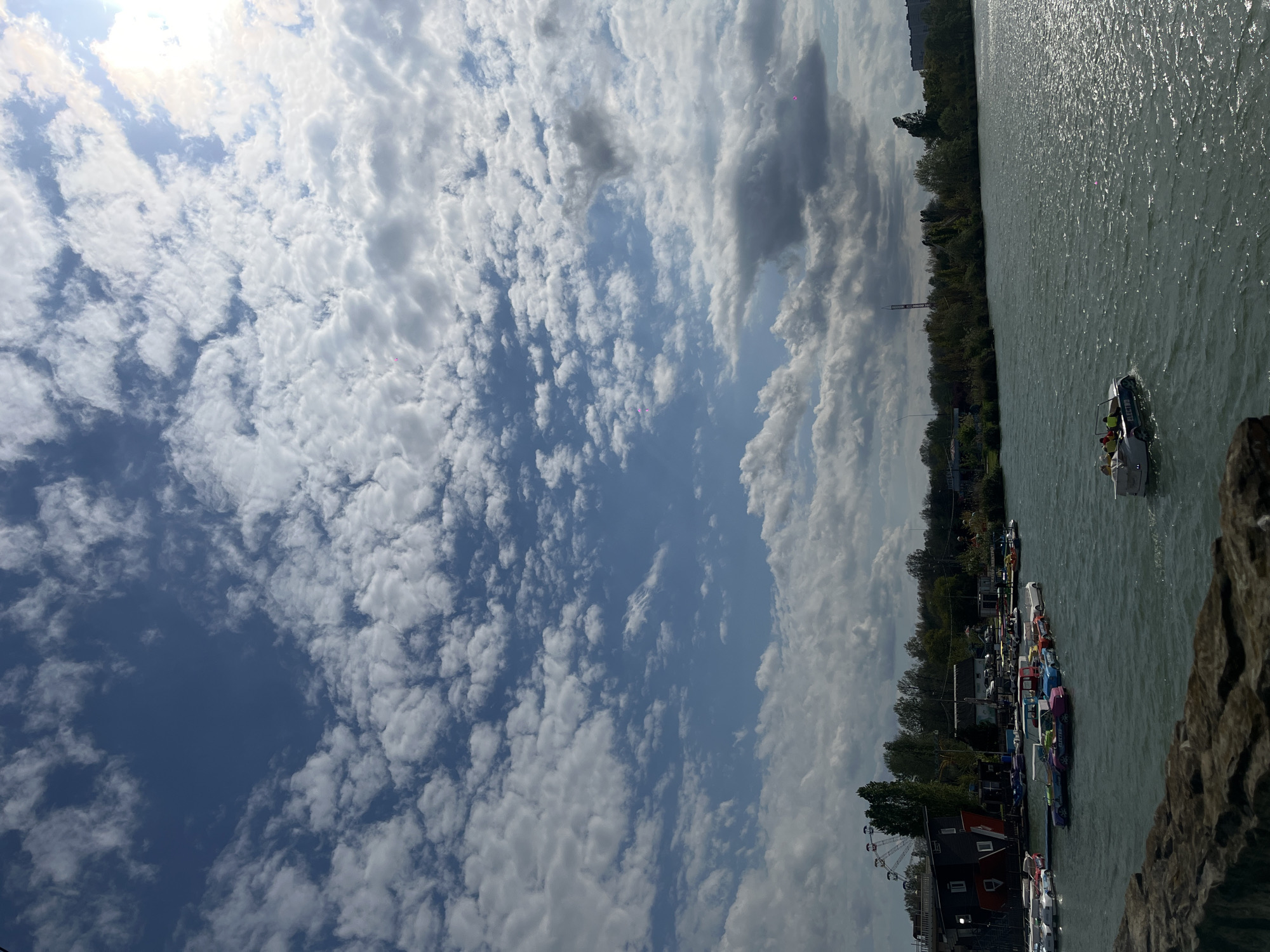 The image shows a waterfront scene in Krasnodar, Russia. A cloudy sky with cumulus formations dominates the left side of the image. On the right, water reflects sunlight, with the shoreline visible in the mid-ground. A variety of paddle boats are docked along the shore. A Ferris wheel is visible in the distance near a building. A small boat is traveling on the water.