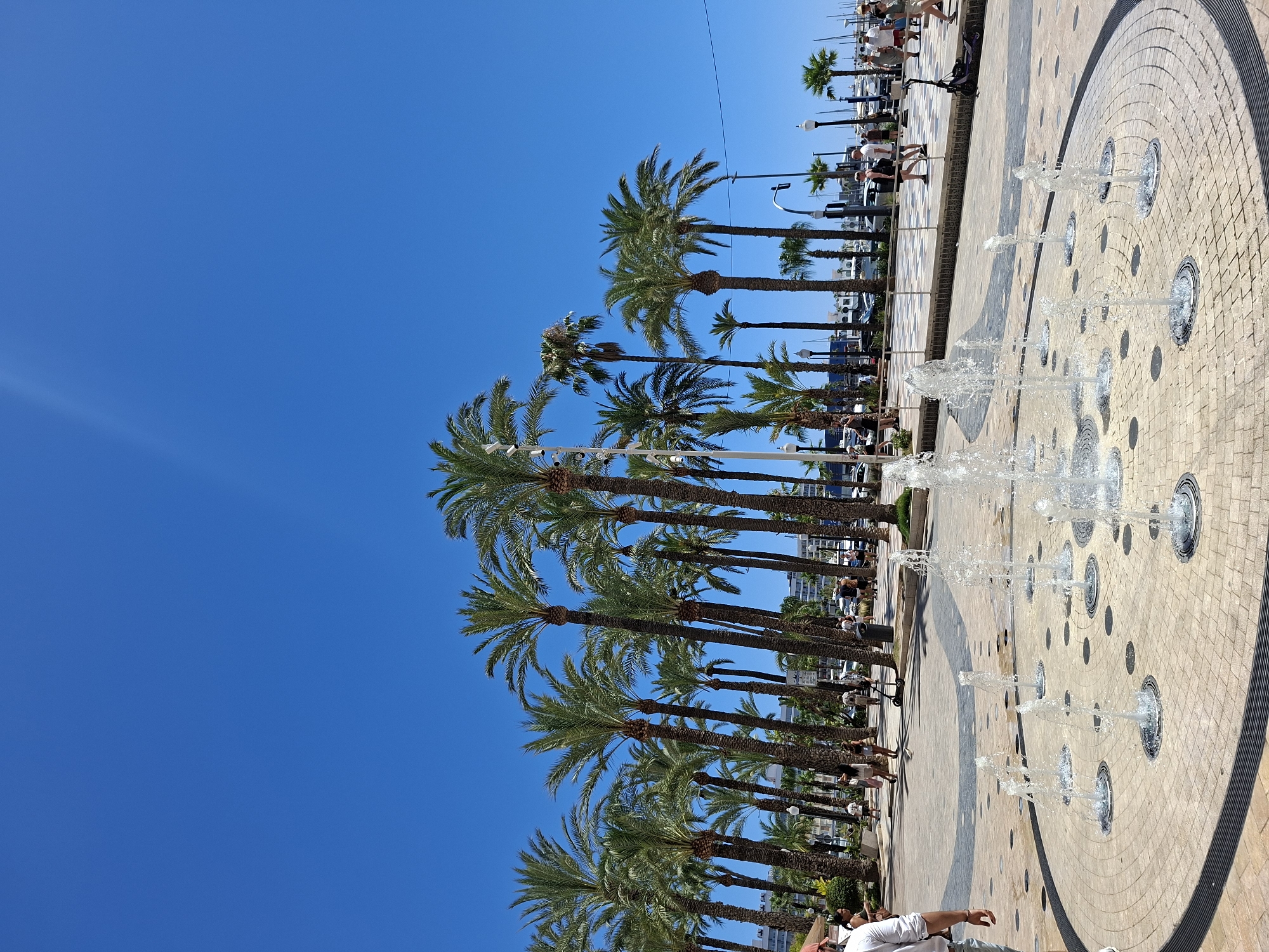 The image depicts a sunny day in Alicante, Spain, likely along the Explanada de España due to the presence of numerous palm trees, a promenade with people walking, and the clear blue sky. In the foreground, a circular water fountain is active, with several jets of water spraying upwards from the pavement. The fountain area is paved with circular patterns of light-colored tiles.
Further back, a wide paved walkway lined with mature palm trees extends horizontally across the frame. People are seen strolling along this walkway, some in casual attire, suggesting a tourist or recreational setting. A few are riding scooters. In the background, buildings and boats in a harbor can be glimpsed through the palm trees, confirming a coastal city location. The overall atmosphere is bright and lively, indicative of a warm, pleasant day.