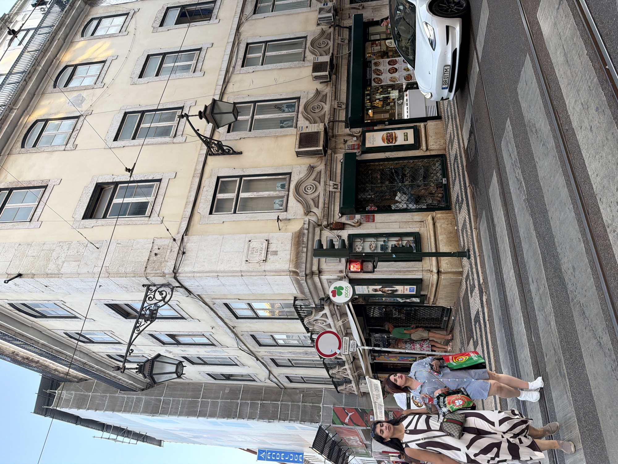 The image depicts a street scene in Lisbon, Portugal, specifically on Rua da Conceição. In the foreground, two women are walking across a zebra crossing, with tram tracks visible on the road. The woman on the left wears a patterned dress and carries a crossbody bag. The woman on the right is in a striped dress and holds several shopping bags, including one with a Portuguese flag design.
Behind the women, a building with cream-colored facade and stone accents lines the street. The building features multiple windows, balconies, and ornate architectural details. Traditional street lamps are affixed to the building's facade. A traffic light is visible on the right side of the frame, showing a red signal.
Several shops are visible at the ground level of the building. One shop has a sign that reads "JOGOS SANTA CASA" and displays colorful items. Another shop has a menu board featuring food items, including a burger. A white electric vehicle, identified by its license plate "BR01TM", is stopped at the crossing. In the background, scaffolding and a large banner are present on another building, suggesting ongoing construction or renovation. The sky above is clear and blue.