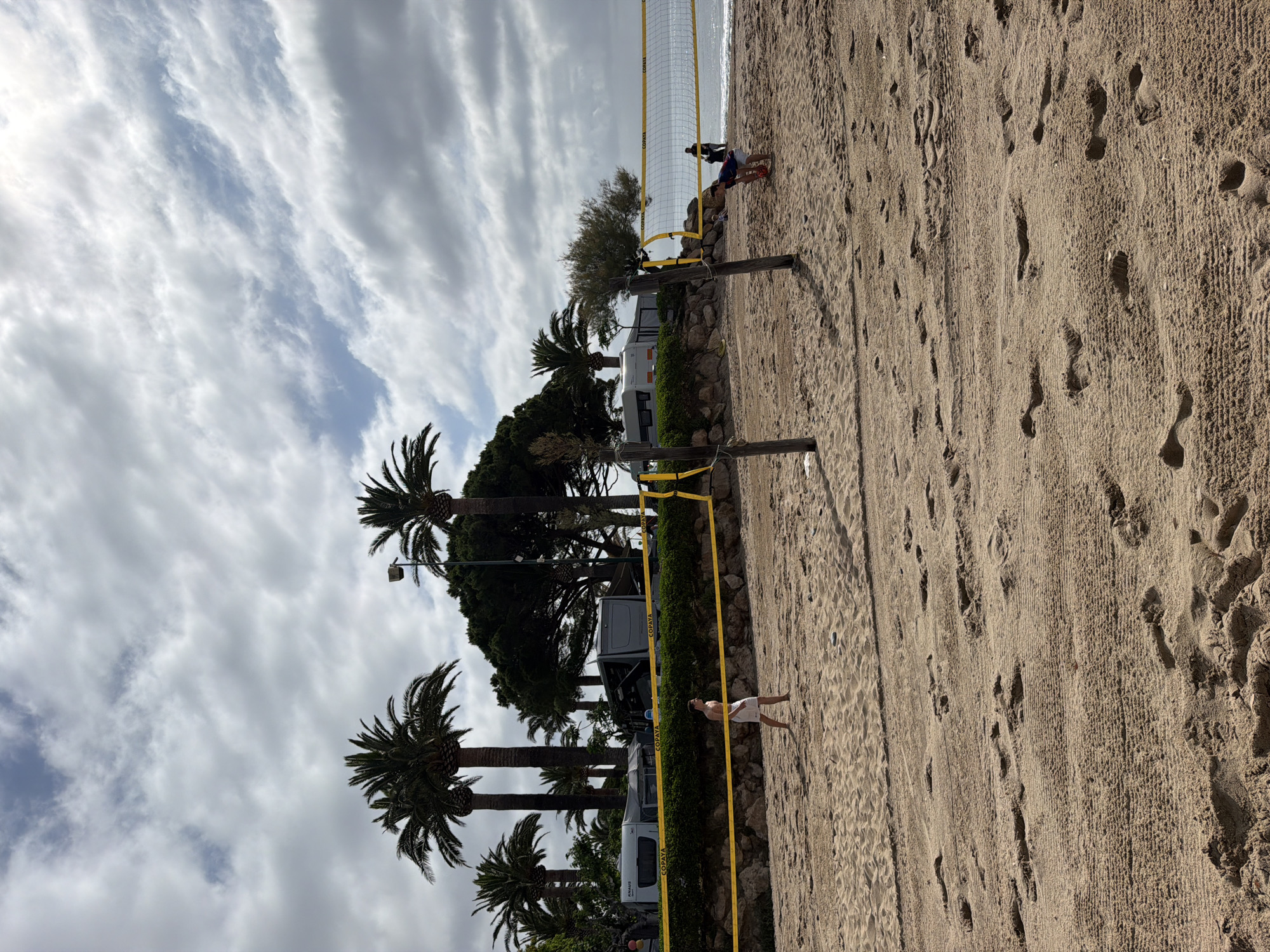This image depicts a beach volleyball court on a sandy beach in Mont-roig del Camp, Spain, under an overcast sky. Two male individuals are present on the court. On the left, a fair-skinned male, shirtless and wearing white shorts, stands barefoot on the sand, facing towards the right side of the court in an active stance. On the right, a second fair-skinned male, wearing a dark shirt and shorts, crouches near the net, holding a red and white volleyball, poised to serve.
The volleyball net, bearing "COPAYA" branding on its yellow tape borders, is stretched between two rough-hewn wooden poles. The sand in the foreground and on the court shows numerous footprints and textured patterns, possibly from beach grooming. In the background, the Mediterranean Sea is visible, appearing calm with a subtle horizon line.
Behind the beach, a low, rocky embankment topped with dense green foliage and smaller, gnarled trees separates the sand from an area containing several parked recreational vehicles (RVs) or caravans. Notable vehicles include a white "KNAUS" branded RV and a darker "COPAYA" branded RV, with other vehicles partially obscured by tall palm trees that punctuate the skyline. A streetlamp is also visible above the foliage. The sky is largely covered by grey stratocumulus clouds, with some lighter patches indicating thinner cloud cover or breaks. Two additional human figures are visible as dark silhouettes on the wet sand near the water's edge in the far background, one standing and one further to the right.