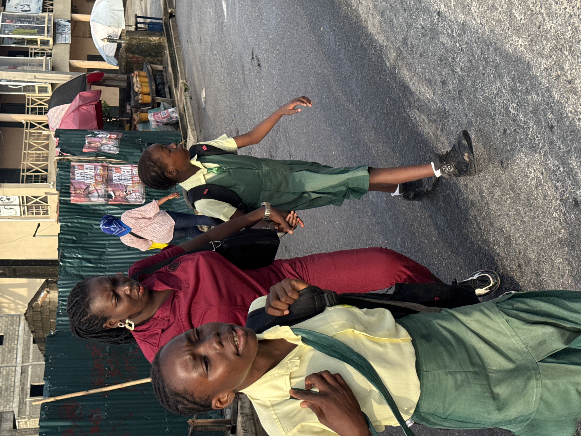 In Ado, Nigeria, on an asphalt road, a woman in a maroon top and a girl in a yellow shirt and green jumper walk beside another girl in a similar school uniform. The woman is wearing large, gold hoop earrings. The younger girl in the yellow shirt walks slightly ahead, looking to her left, with a black backpack. The girl in the green jumper walks to her right, holding the hand of the woman. The woman is also carrying a black bag over her shoulder. In the background, there is a green corrugated metal fence with posters and some pink and black umbrellas. Further back, there are buildings, one with balconies and windows, and a white umbrella. A vendor appears to be selling produce from buckets. The scene depicts a typical street in Ado, Nigeria, with individuals on their daily commute or journey.
