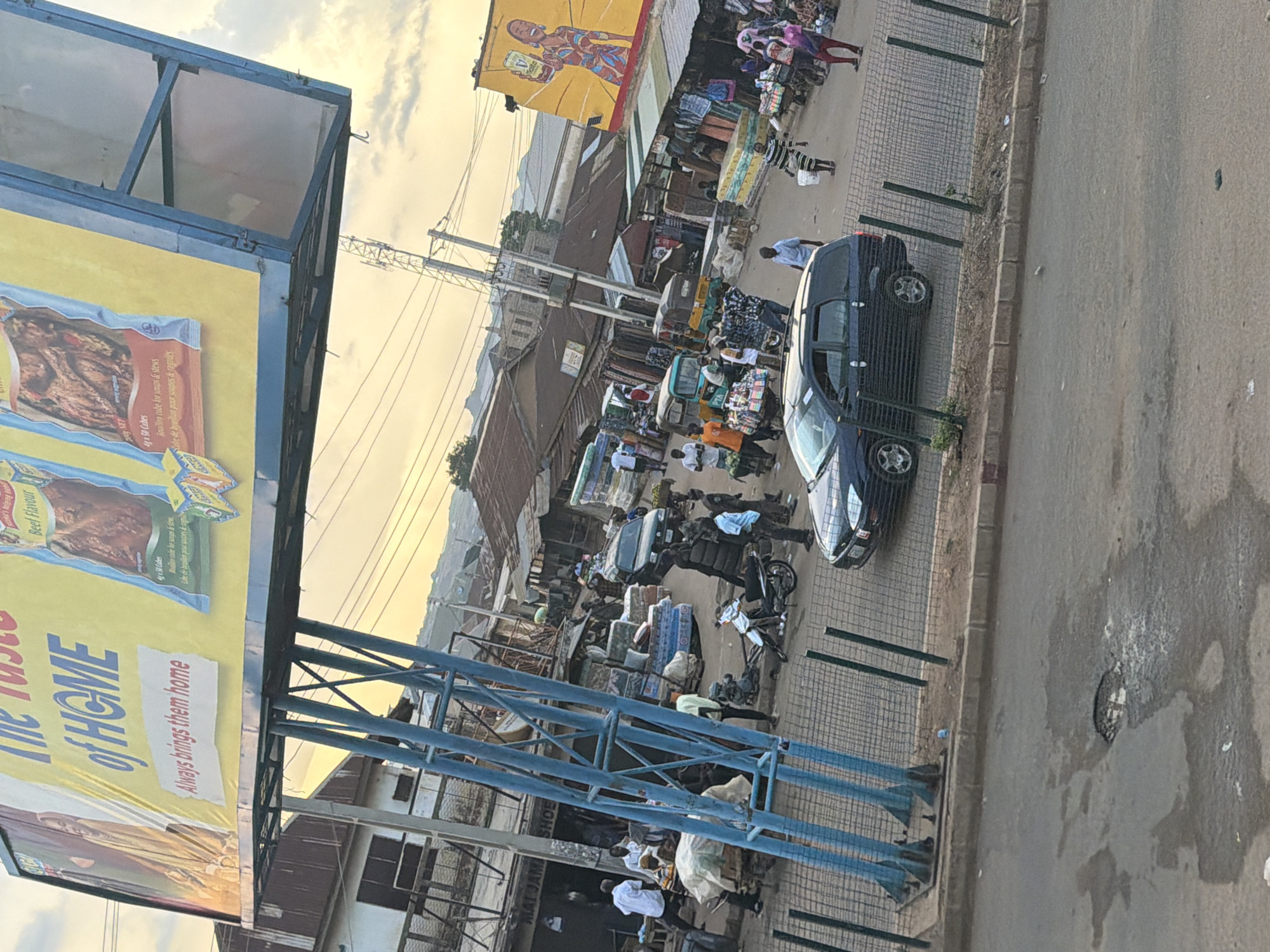 The image depicts a bustling marketplace scene in Bukuru, Nigeria, with a large billboard prominently displayed overhead. The billboard features an advertisement with the text "The Taste of HOME" and images of food products, likely bouillon cubes. Below the billboard, a busy street is filled with people, stalls, and vehicles.
In the foreground, a black station wagon is parked, and a motorcycle is leaning on its side. A group of people are gathered around stalls displaying various goods, including mattresses and what appear to be textiles. Several individuals are seen walking and interacting within the market. In the background, more market stalls and buildings line the street, with a general atmosphere of commerce and daily life. The sky above is overcast, suggesting either early morning or late afternoon light. A prominent blue metal structure supporting the billboard stands in the middle ground. The road surface shows signs of wear and tear, including a large pothole filled with water.