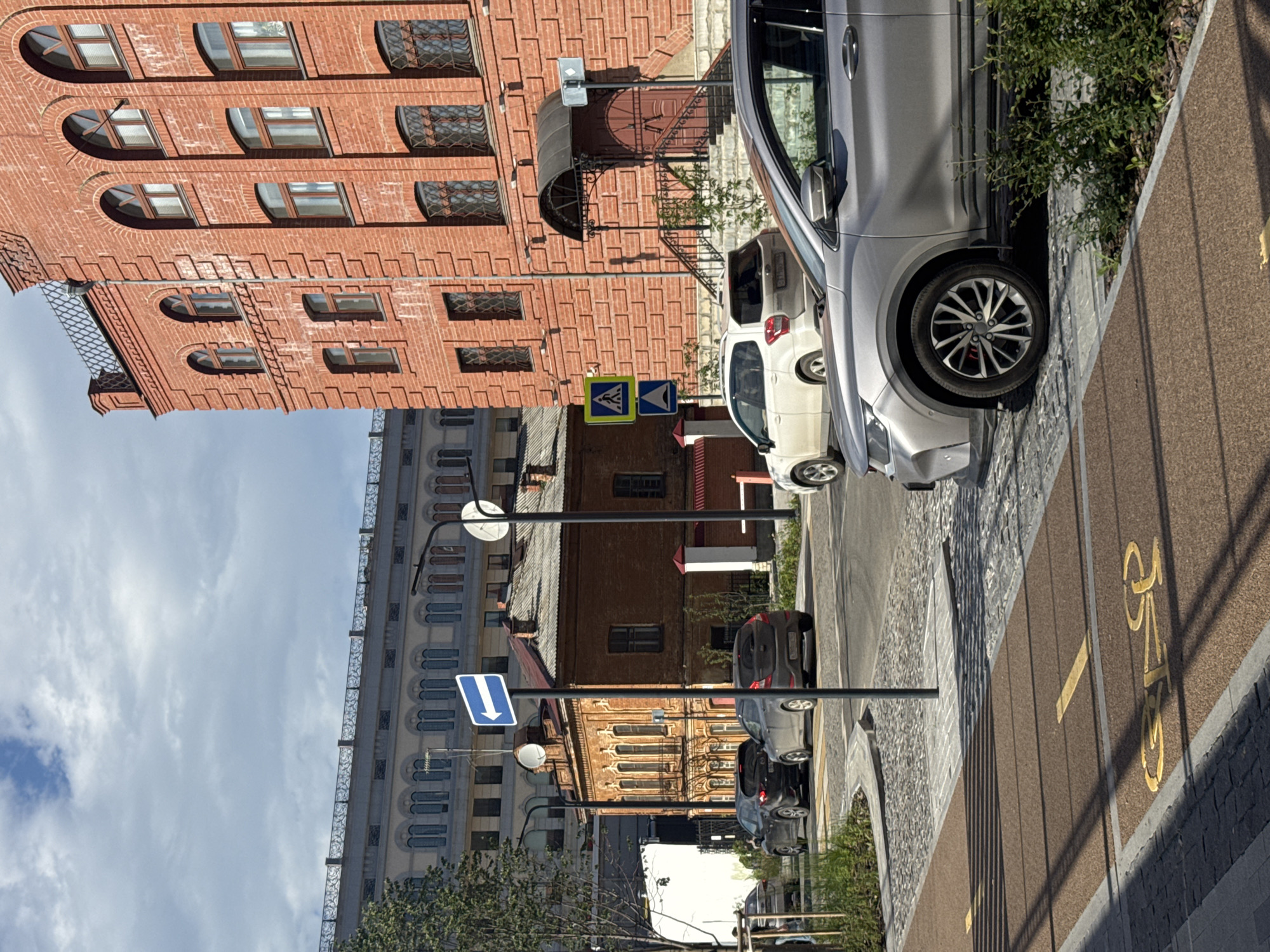 The image shows a street scene in Yekaterinburg, Russia. The perspective is from an elevated viewpoint, looking down at the street. The time of day appears to be daytime, with bright sunlight casting shadows.
The scene is dominated by several buildings. On the left, a tall, modern building with numerous windows stretches upwards. Adjacent to it is an older, red brick building with arched windows and decorative architectural elements. Further down the street, another brick building with a more classical facade is visible.
Several cars are parked along the street. A light-colored SUV is prominently featured in the foreground. A dark-colored hatchback and a few other vehicles are also visible further down. There are traffic signs, including a pedestrian crossing sign and a directional arrow sign. A bicycle lane is marked on the pavement with yellow paint.
The overall impression is a city street with a mix of architectural styles and urban activity. The weather appears clear and sunny. There is no visible text in the image.