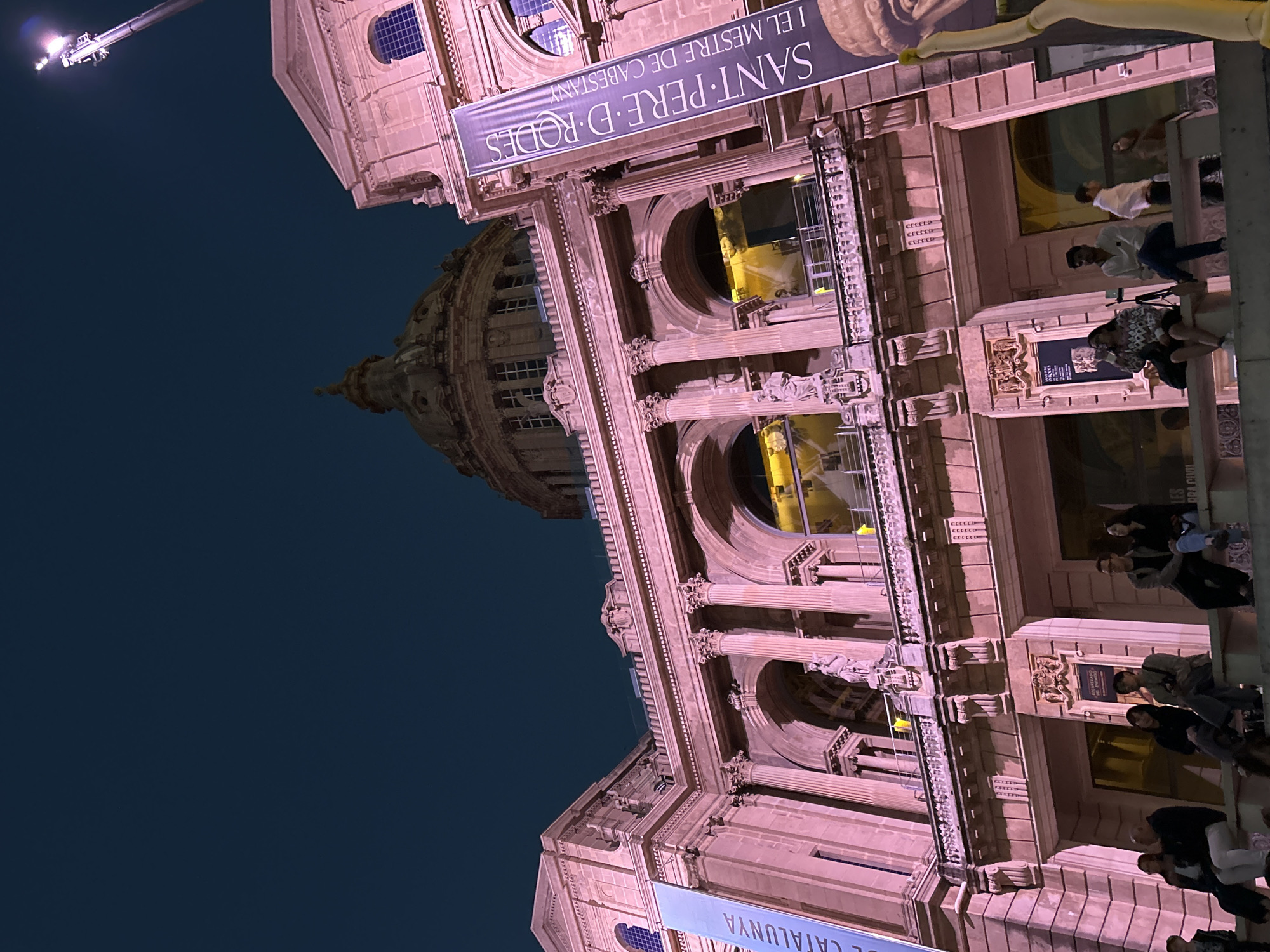 A grand, classical-style building, likely a museum or public institution, is captured at night from a low-angle perspective. The elaborate facade features columns, arches, and detailed stonework, with a large dome partially visible in the background. The building is bathed in a warm, pinkish artificial light, contrasting sharply with the deep dark blue of the night sky.
On the lower levels of the building, a group of people is visible. Some individuals are standing, while others are sitting on what appear to be wide steps or a terrace. They are casually dressed and appear to be gathered informally, possibly waiting for an event or simply enjoying the evening atmosphere.
In the upper left corner, a large crane equipped with bright lights extends upwards, suggesting either construction, maintenance, or specialized event lighting for the building. The sky is clear, indicating fair weather.
Several banners are affixed to the building. One prominent banner displays the text "SANT PERE D'RODES" (Saint Peter of Rodes) and "I EL MESTRE DE CABESTANY" (and the Master of Cabestany), likely referencing an exhibition. Another partially visible banner includes the words "BRA CIVIL" (Civil War) and "L CATALUNYA" (Catalonia), further hinting at a historical or cultural theme, possibly "Civil War in Catalonia." Given the context of Barcelona, Spain, this impressive structure is very likely the National Art Museum of Catalonia (MNAC) or a similar cultural landmark.